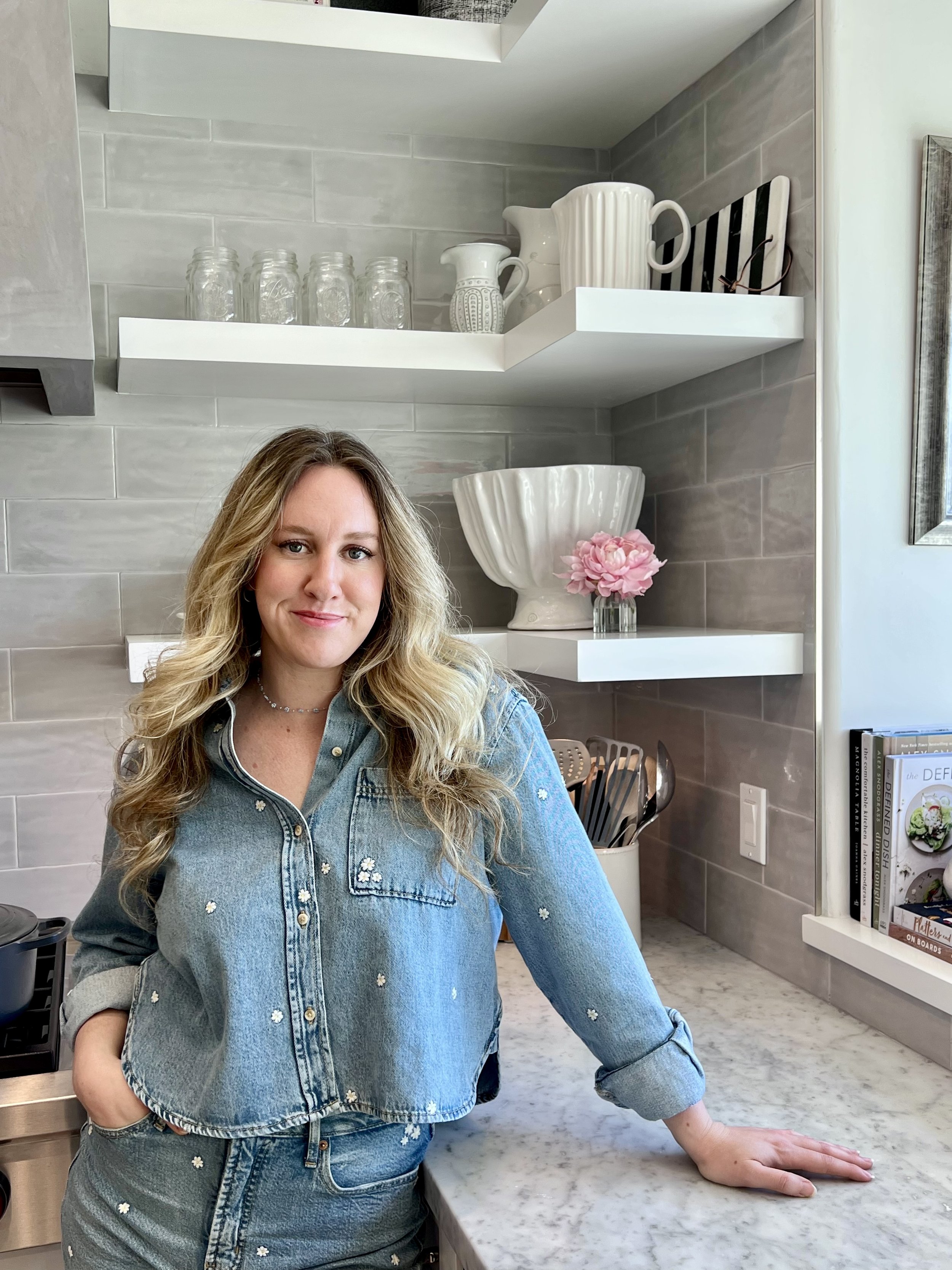 A woman with long blonde hair and a denim outfit stands in a modern kitchen with grey tile backsplash, open white shelves with glassware and decorative objects, and a marble countertop.
