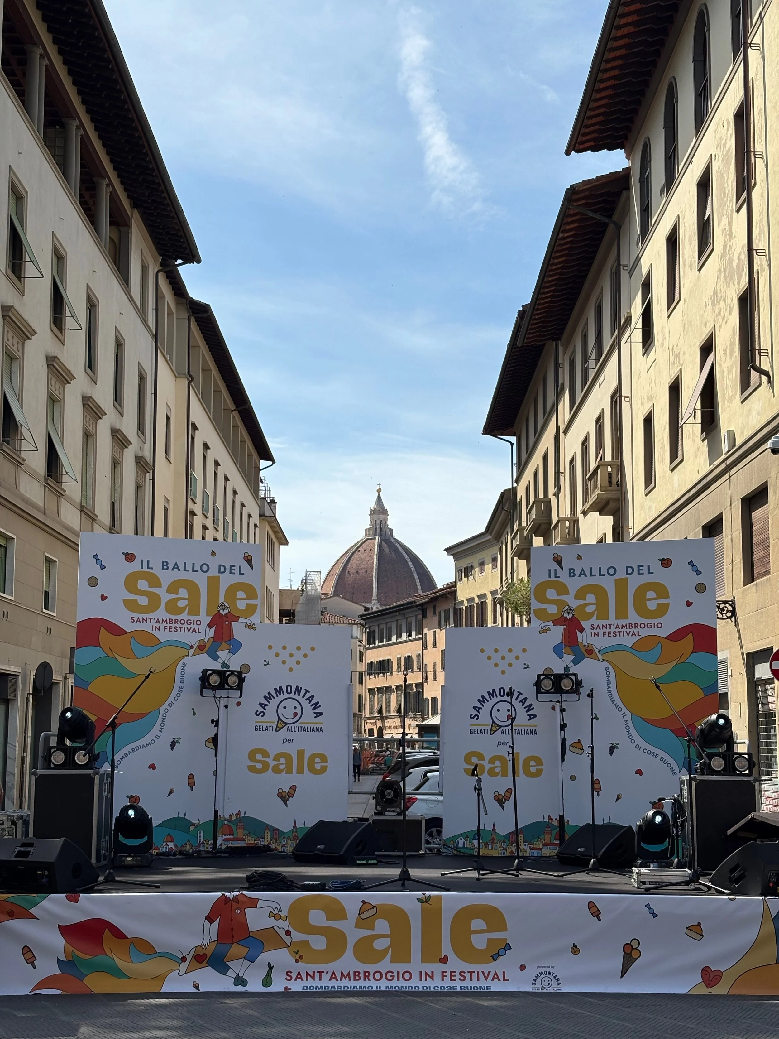 Stage set up for a festival event in Florence, Italy with multi-story buildings on either side and a dome building in the background, featuring colorful banners, lights, and sound equipment.