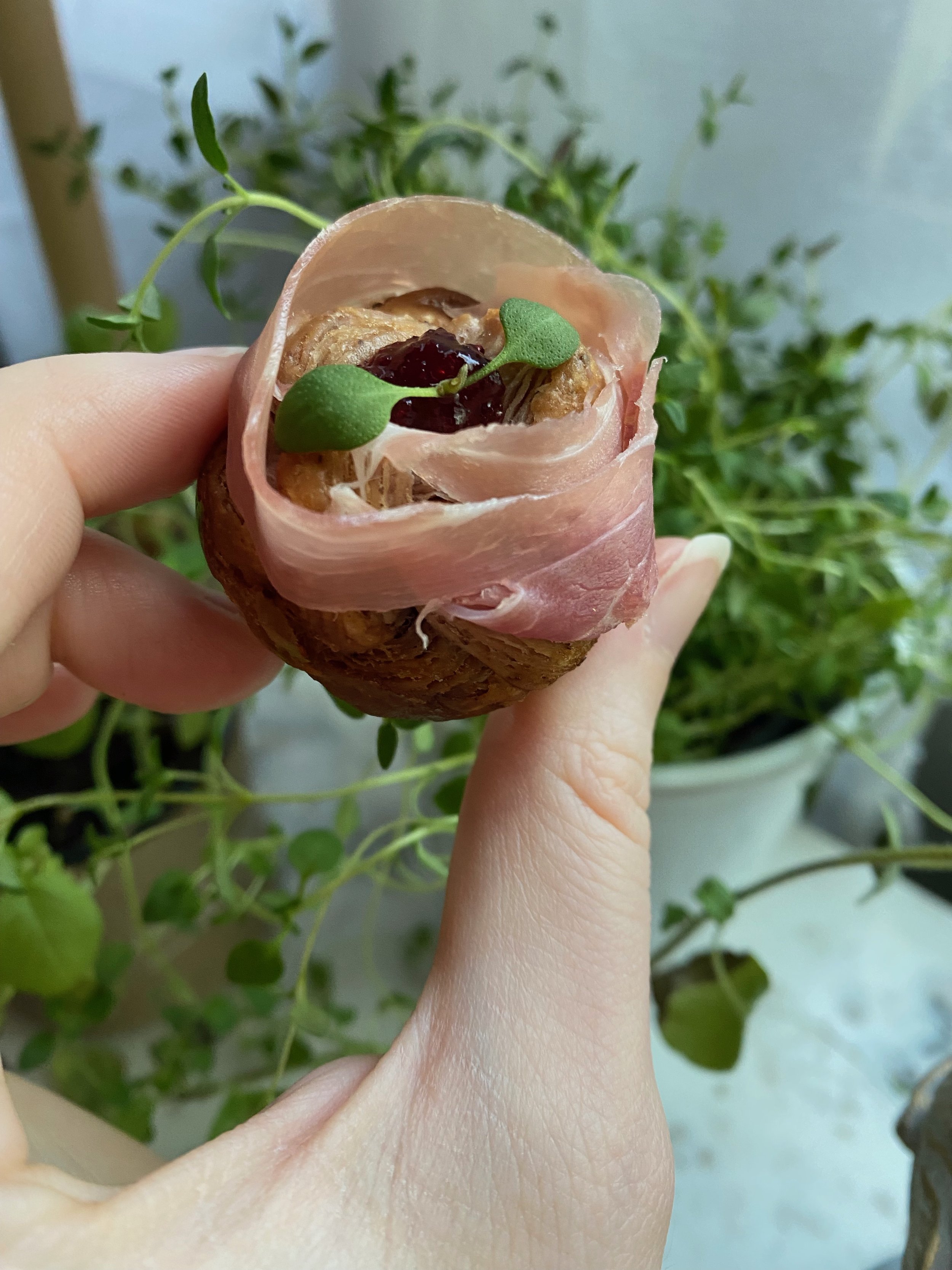A hand holding a blue corn croissant with prosciutto, topped with a small green leaf and a dollop of red jam, with green plants in the background.