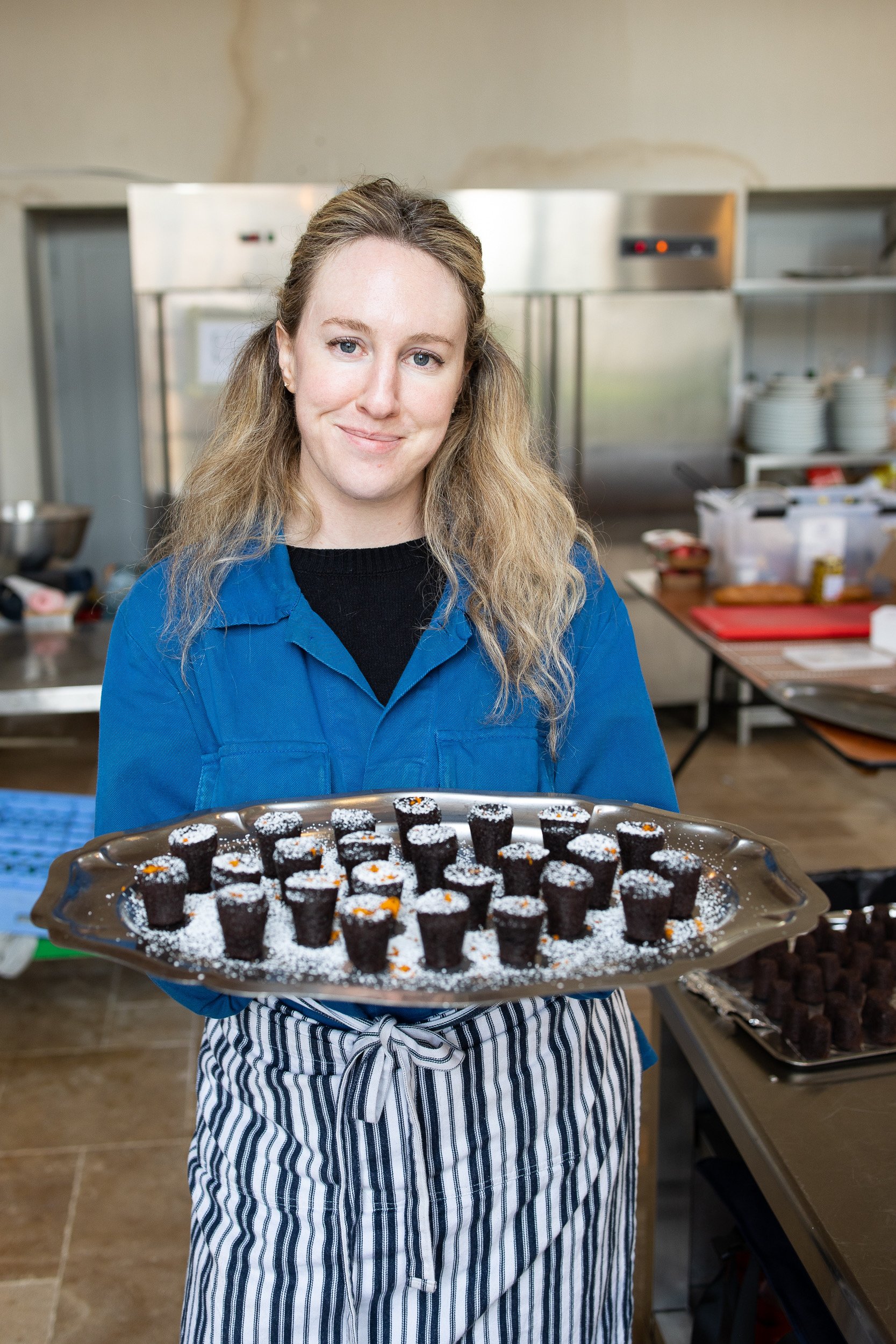A woman with long, wavy blonde hair, wearing a blue uniform and striped apron, holding a tray of small chocolate desserts decorated with powdered sugar and orange zest, in a commercial kitchen.