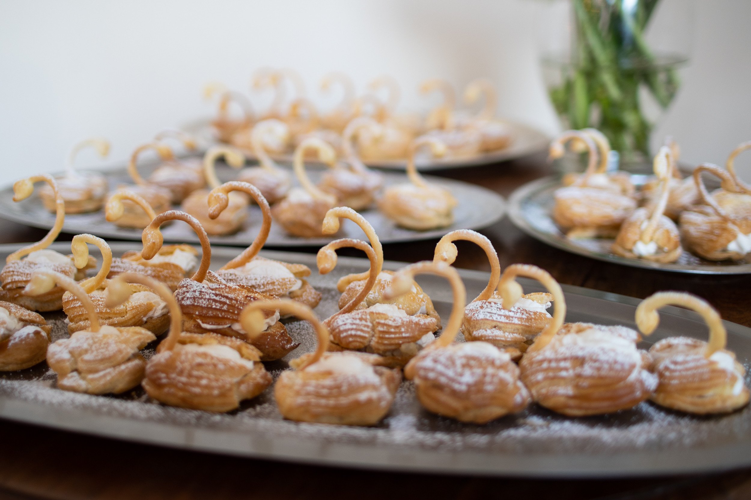 Trays of cream puffs garnished with powdered sugar and decorative curls of pastry, with a vase of green tulips in the background.