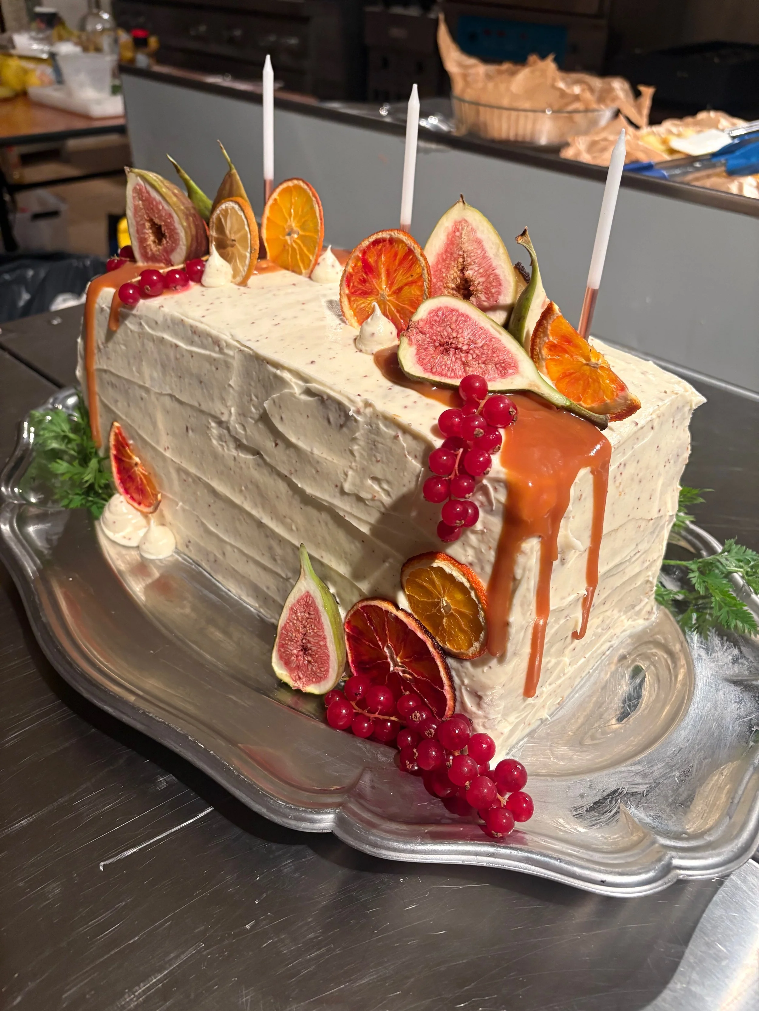 A large rectangular layered cake decorated with figs, dried orange slices, red currants, and caramel drizzle, placed on a silver platter with green garnish.
