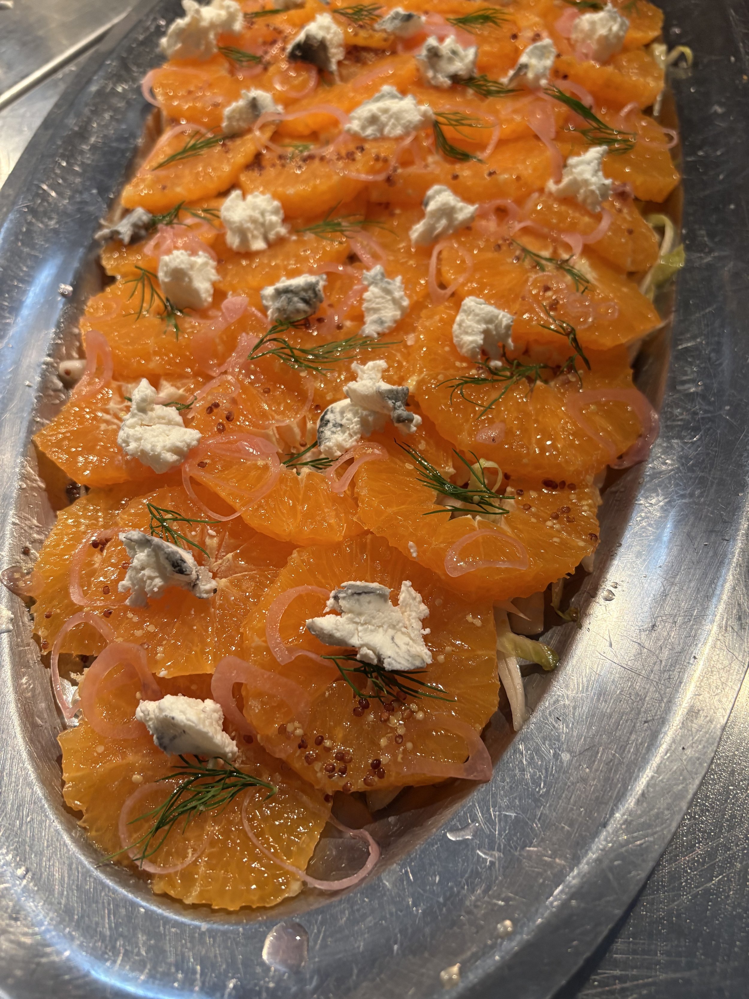 A dish with sliced oranges, pink onions, dill, goat cheese, and mustard seeds on a silver platter.