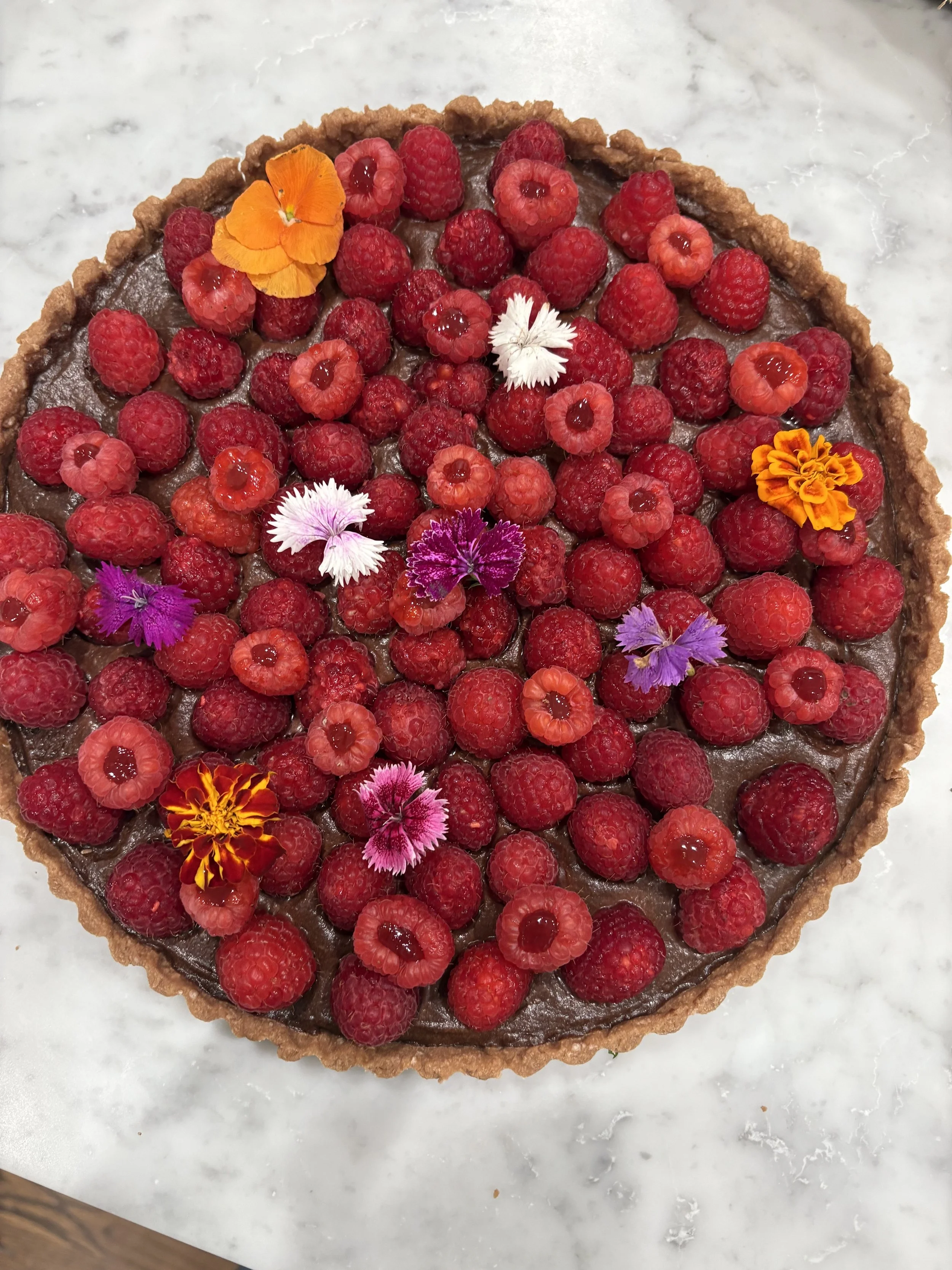 A round chocolate tart topped with raspberries, edible flowers, and dollops of red jam, set on a white marble surface.