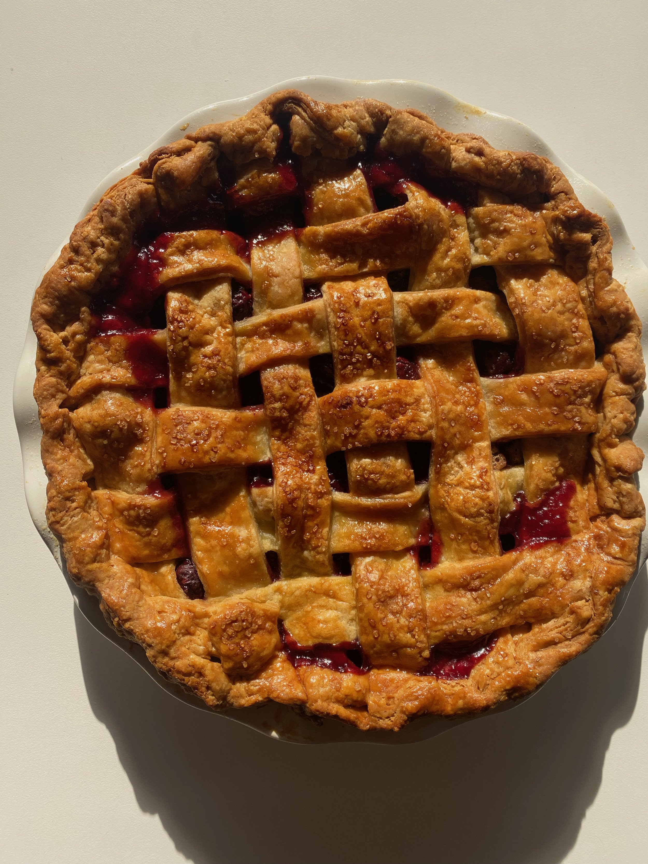 A homemade cherry lattice pie on a white plate with a crust border and a glazed top, set on a white surface with shadow.