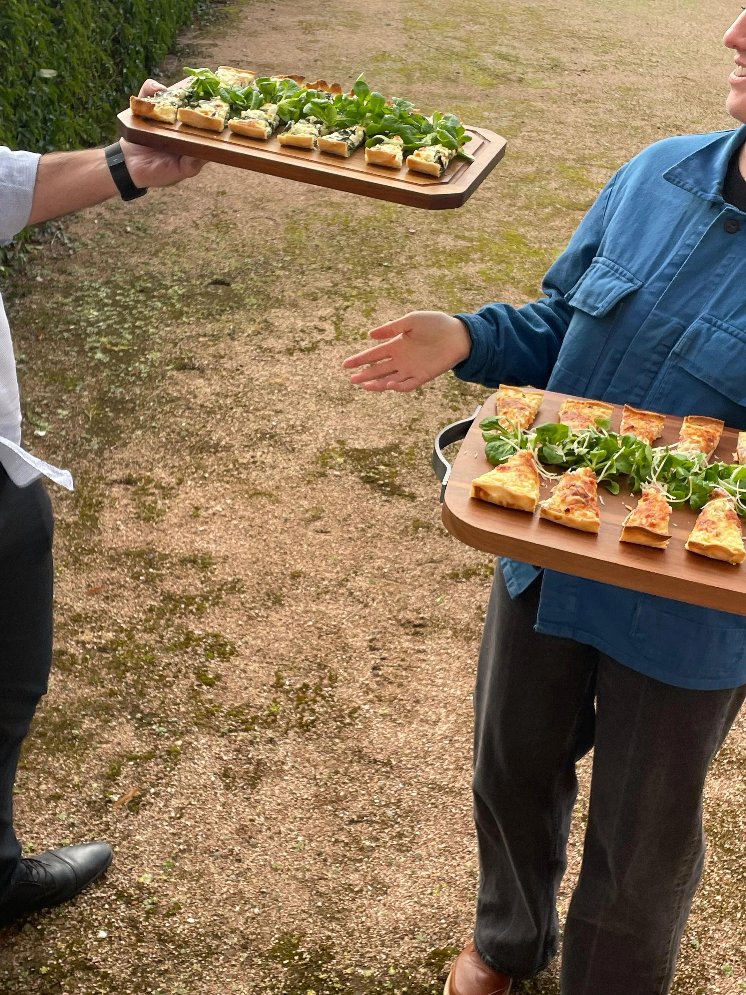 Two people are exchanging trays with assorted quiche slices. The scene appears to be at an outdoor gathering.