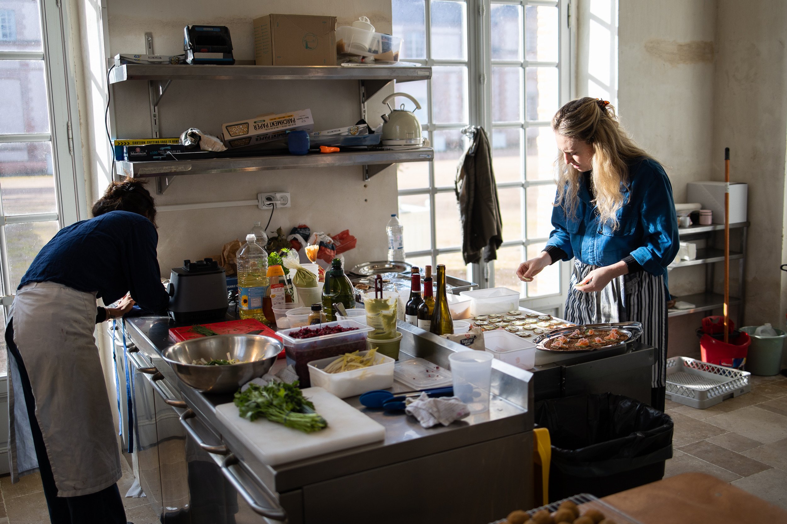 Two women preparing food in an industrial kitchen with large windows, various ingredients, and kitchen supplies on the counters.