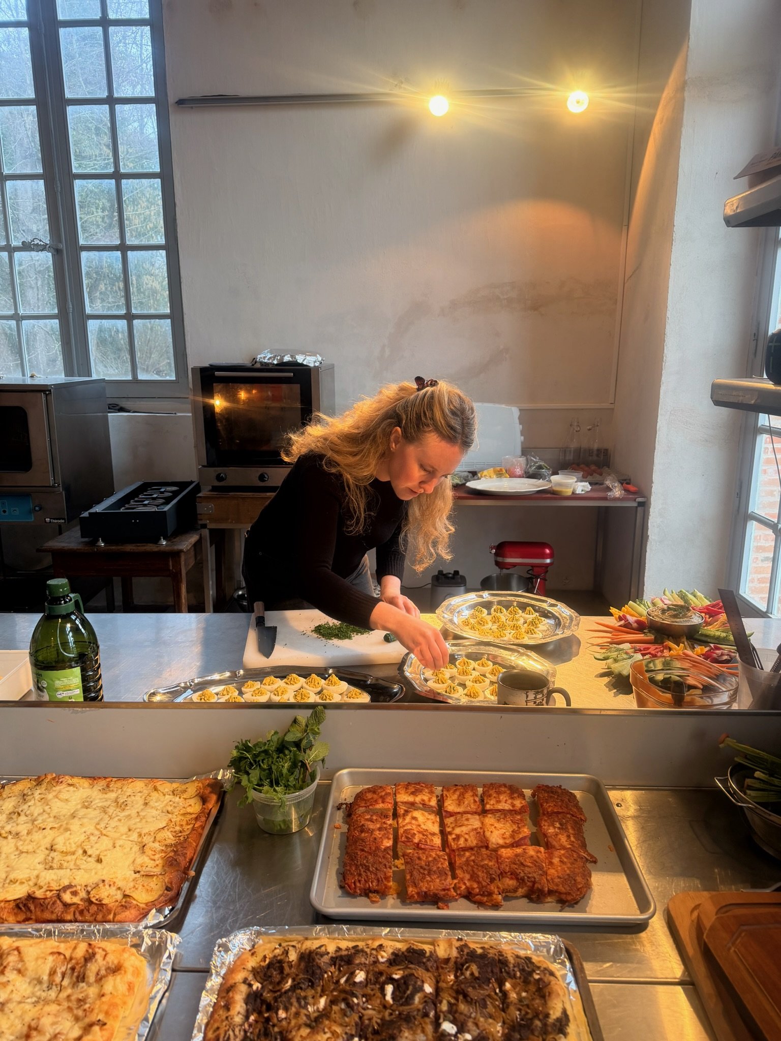 A woman preparing food in a kitchen with various dishes, including pizzas and snacks, on a counter in front of her.