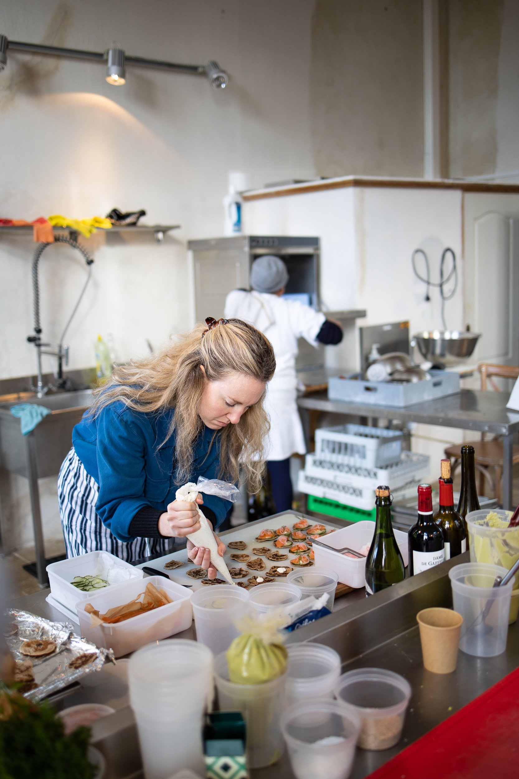 Women preparing food in a kitchen with various ingredients and bottles on the counter, and a person working in the background.
