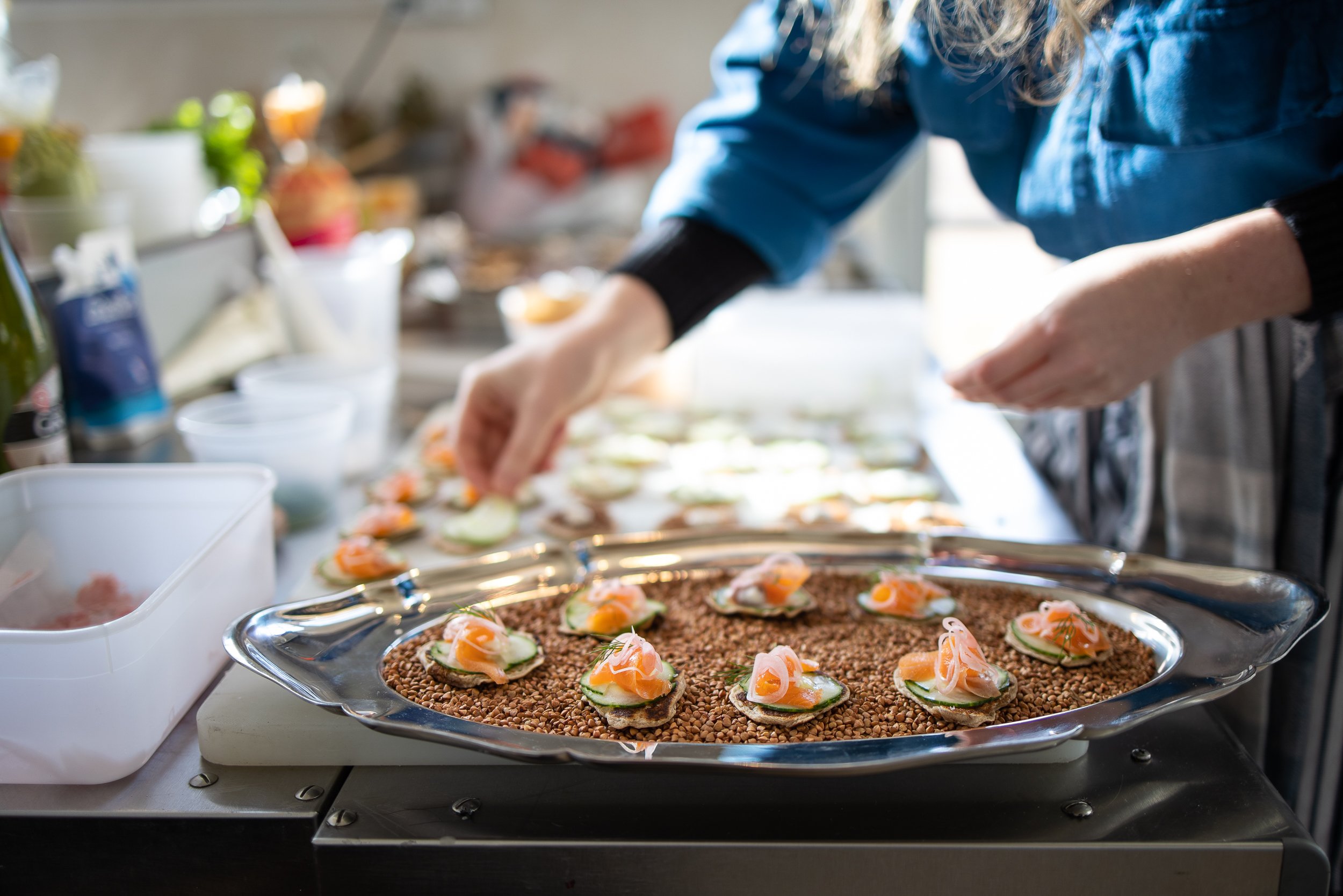 A person preparing small appetizers with smoked salmon, cucumber, and creme fraiche on a large platter lined with buckwheat in a kitchen.