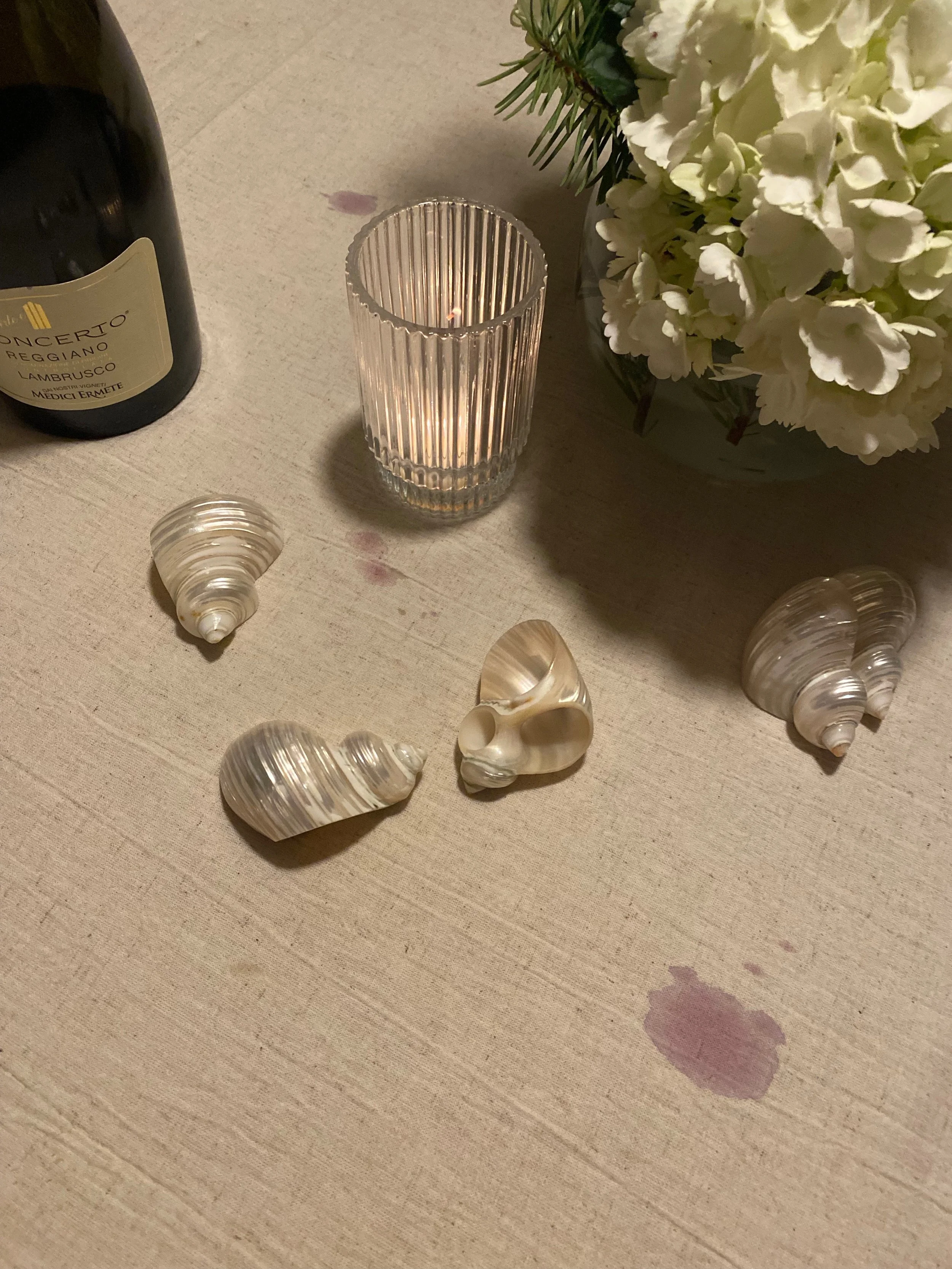 A table with a bottle of red wine, a lit ribbed glass candle holder, a bouquet of white flowers in a black vase, and four decorative seashells on a beige tablecloth.