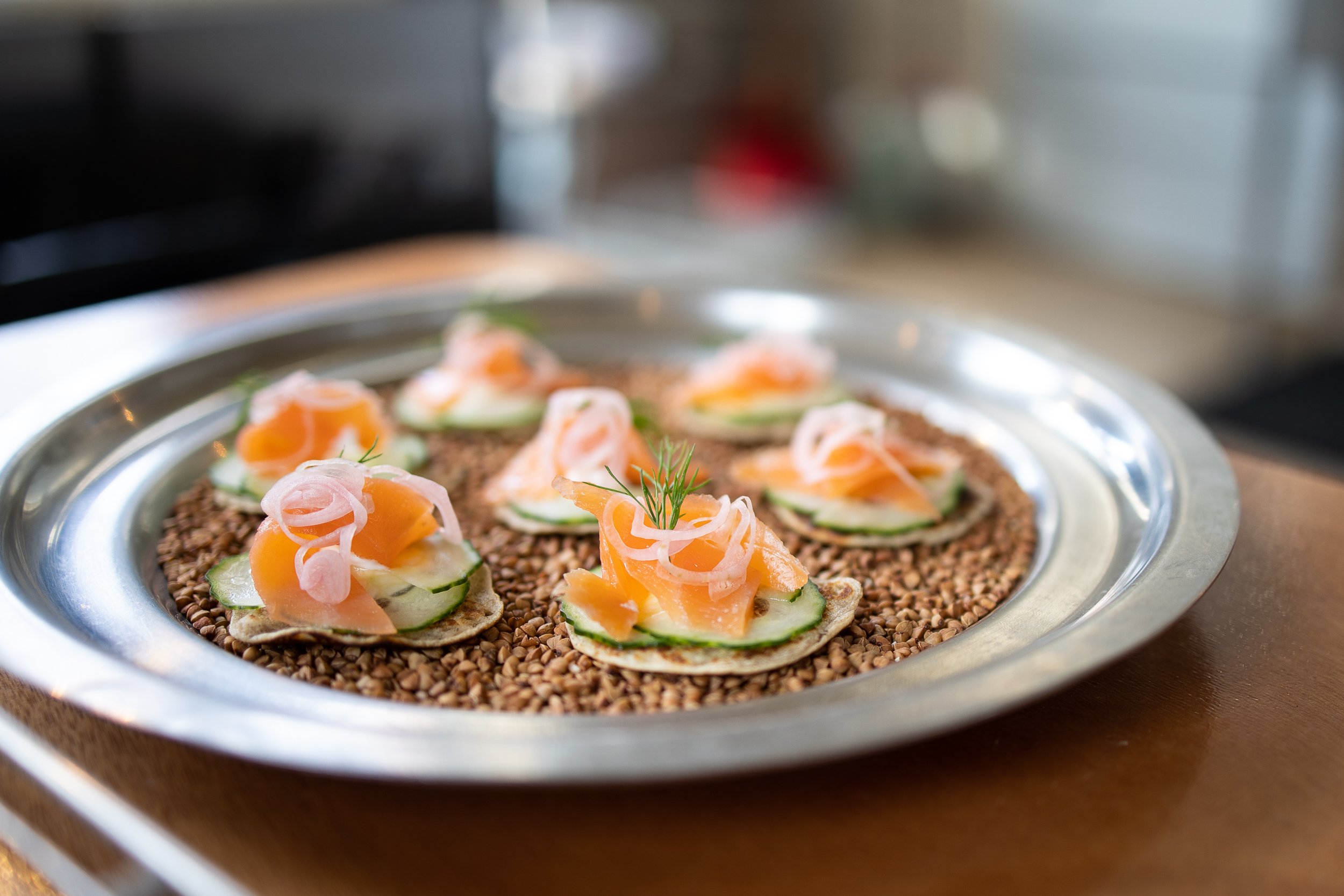 Small appetizers on a silver tray, consisting of buckwheat blinis topped with cucumber slices, smoked salmon, and thinly sliced onions, garnished with a sprig of dill, placed on a bed of buckwheat.
