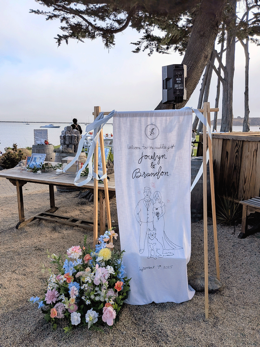 A wedding sign with an illustration of a bride and groom with a dog, on a beachside setup. The sign reads "Welcome to the wedding of Jocelyn & Brandon," dated September 12, 2025. There are flowers in front of the sign and a table with decor and gifts in the background, near the water.
