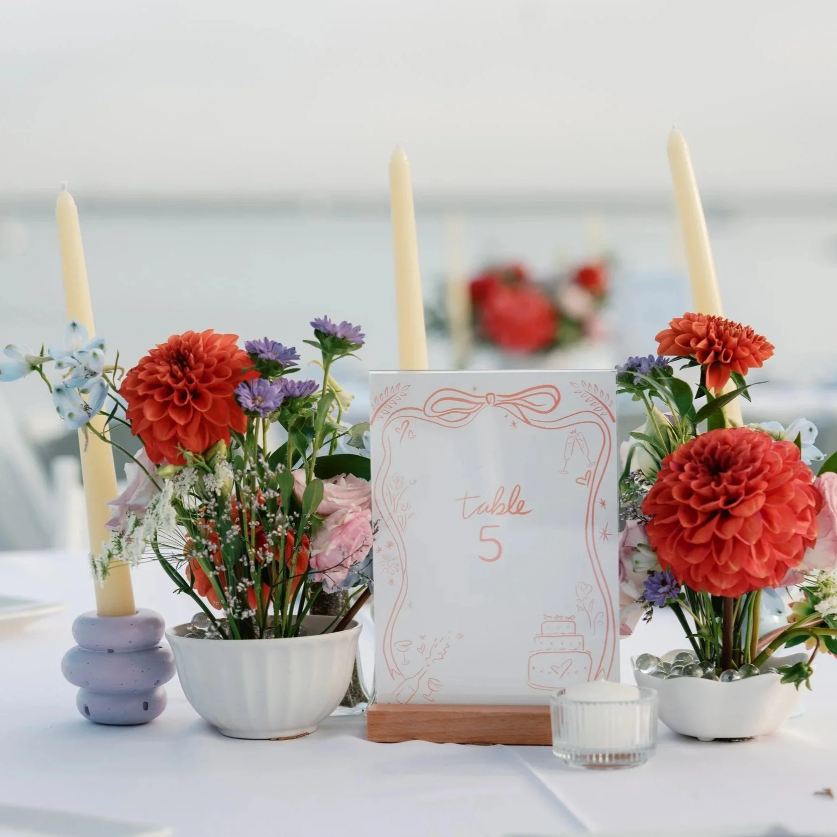 Arrangement of orange, purple, and pink flowers with candles and a sign that says "table 5" at a wedding or event by the water.