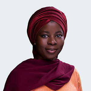 A smiling woman with dark skin wearing a pink headscarf, white blouse, light pink vest, and a wristwatch.