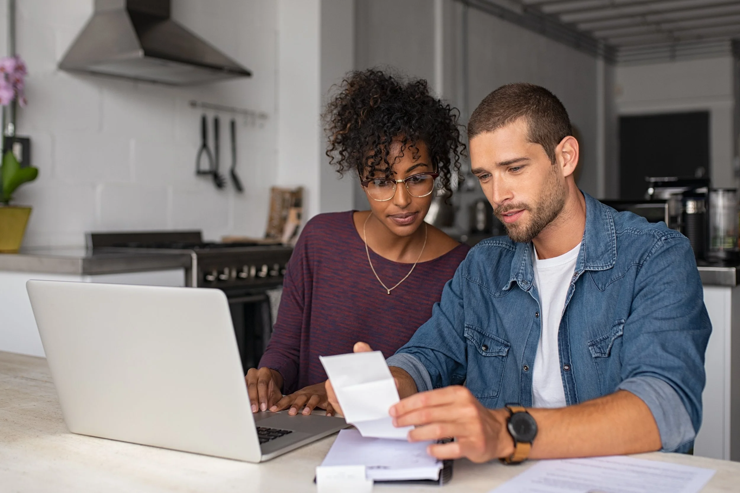 Two people, a woman and a man, looking at a receipt together while sitting at a kitchen table with a laptop and documents.