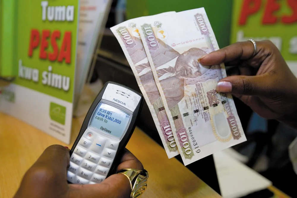 A person is handing over a 100,000 Kenyan shilling bill to another person who is using a mobile phone that shows a cash withdrawal notification of 23,000 KSH. In the background, there is a green book with the title 'Tuma Pesa kwa Simu' on it.