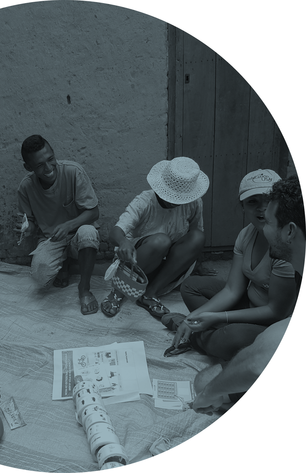 Four people sitting on the ground, engaging in a discussion or activity involving printed materials, pictures, and a roll of stickers, outdoors against a stone wall with a wooden door.