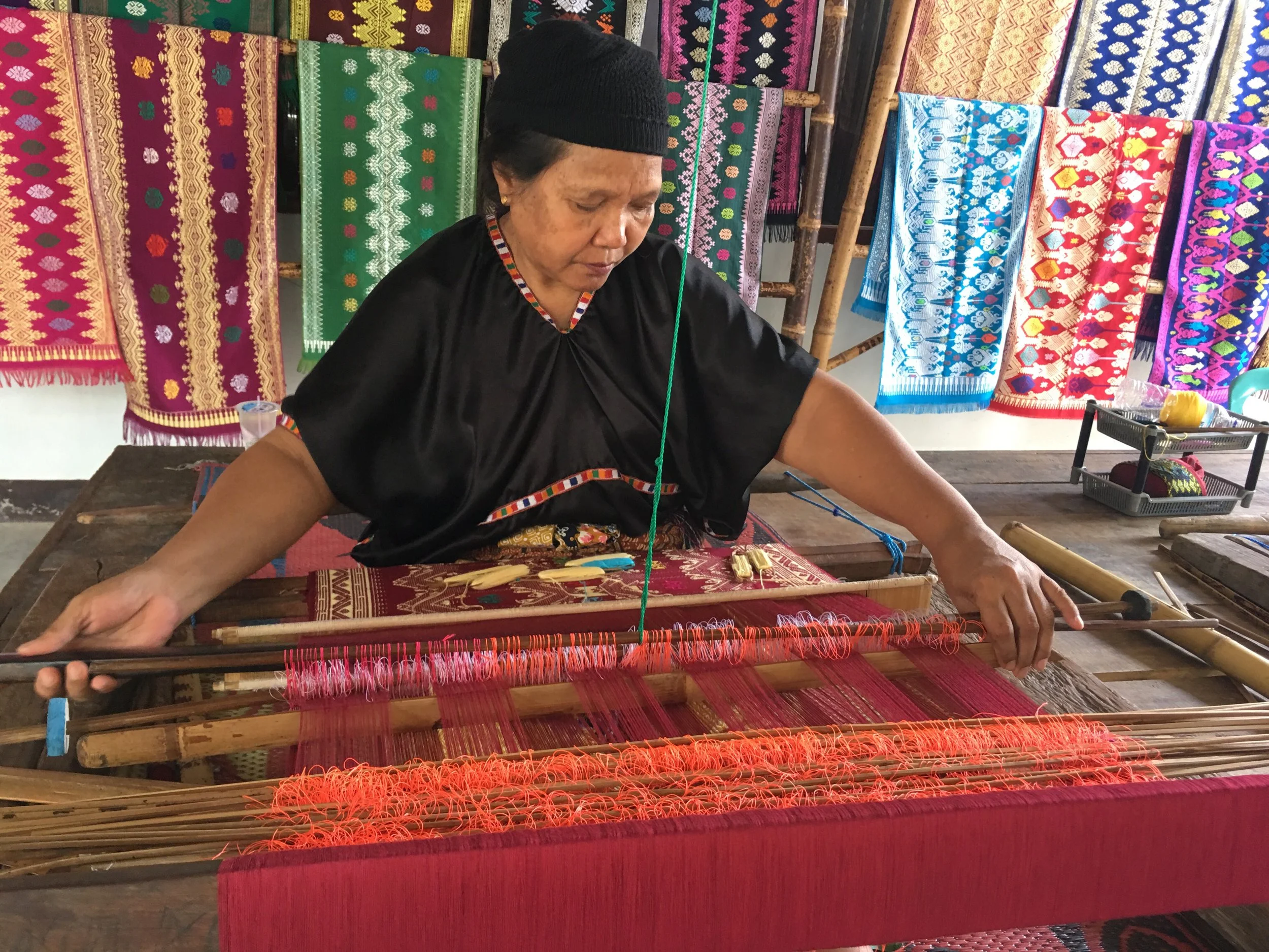 An older woman in black traditional clothing and a black headscarf weaves colorful fabric on a wooden loom. Behind her, vibrant, patterned textiles hang on the wall.