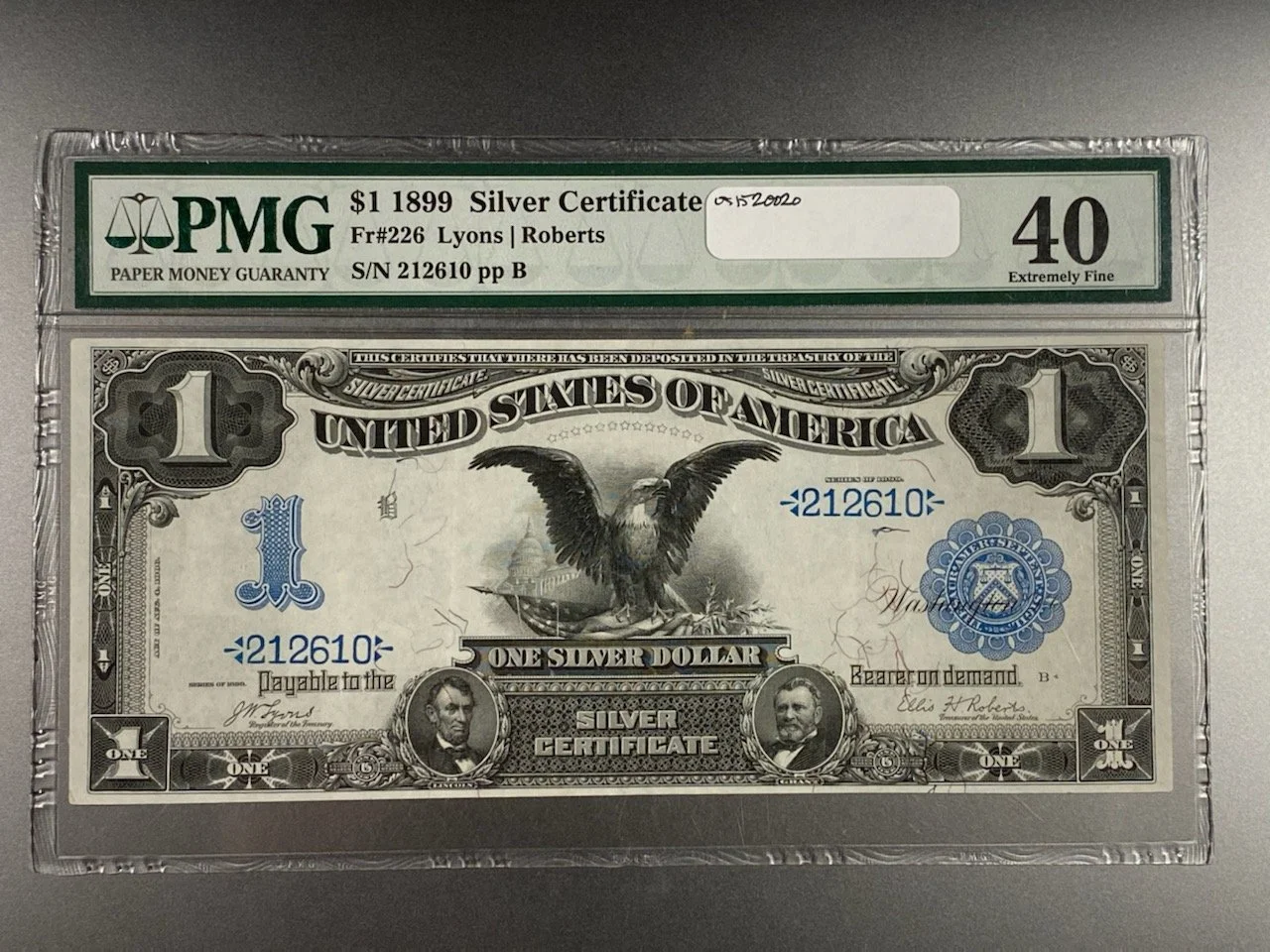 An 1899 Silver Certificate US one-dollar bill encased in a plastic holder with a label from PMG grading it 40, extremely fine. The bill features an eagle with a shield on its chest, holding a branch and arrows, with portraits of two men at the bottom