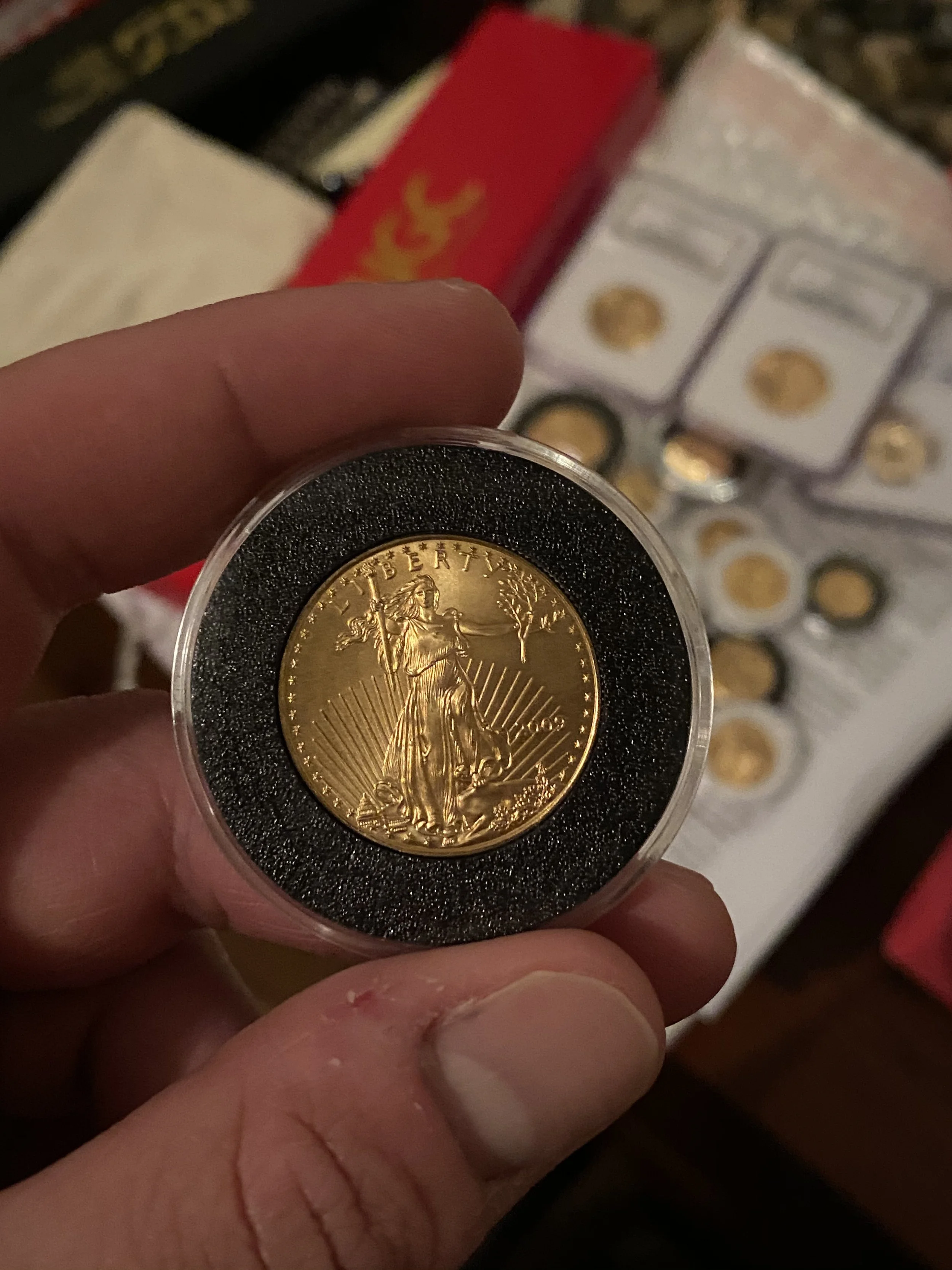 Close-up of a gold coin in a protective case, with a background of more coins and boxes. The coin features the image of Lady Liberty holding a torch, with the word 'LIBERTY' at the top.