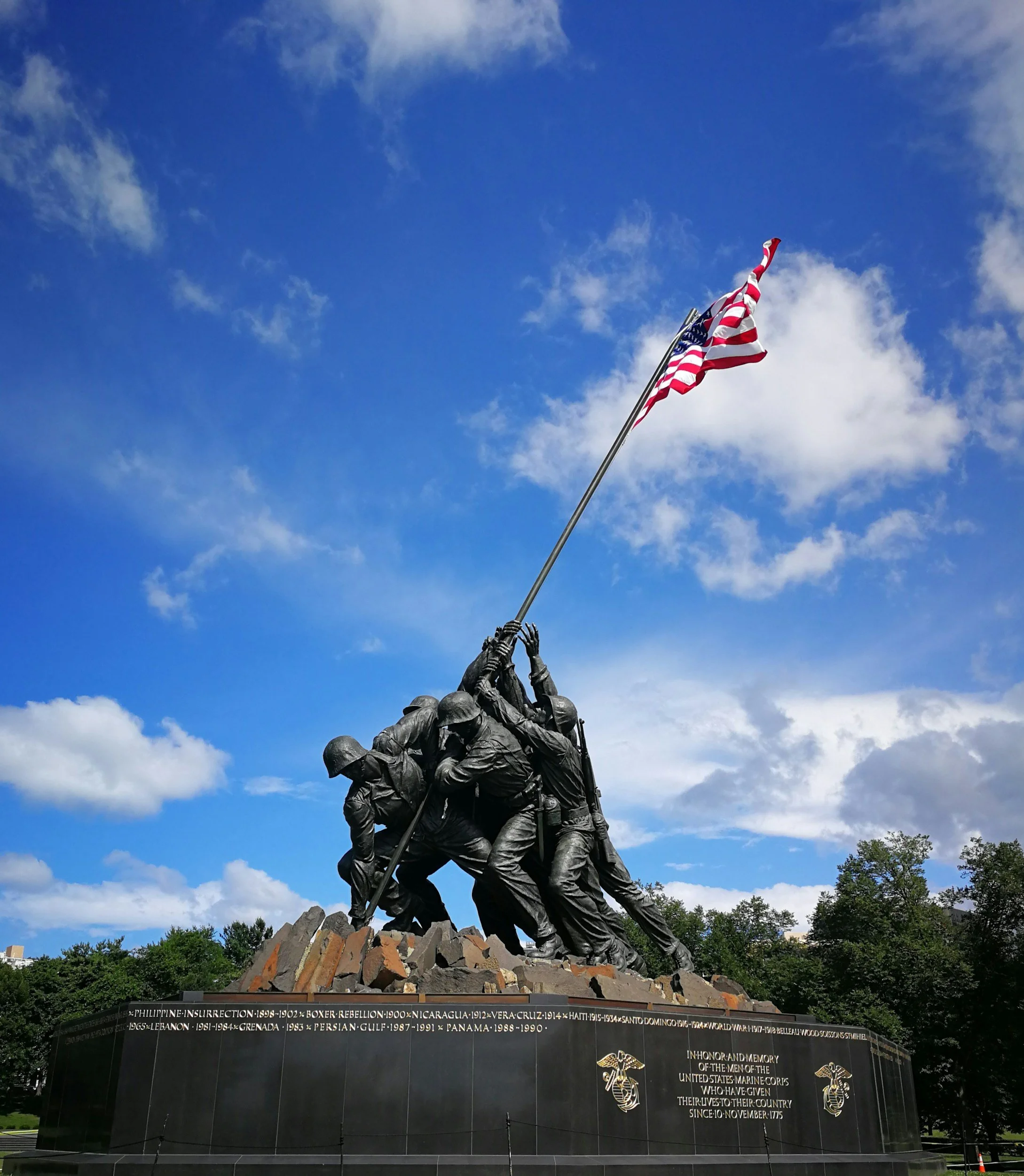 Photograph of the Marine Corps War Memorial featuring soldiers raising an American flag against a partly cloudy sky with green trees at the base.