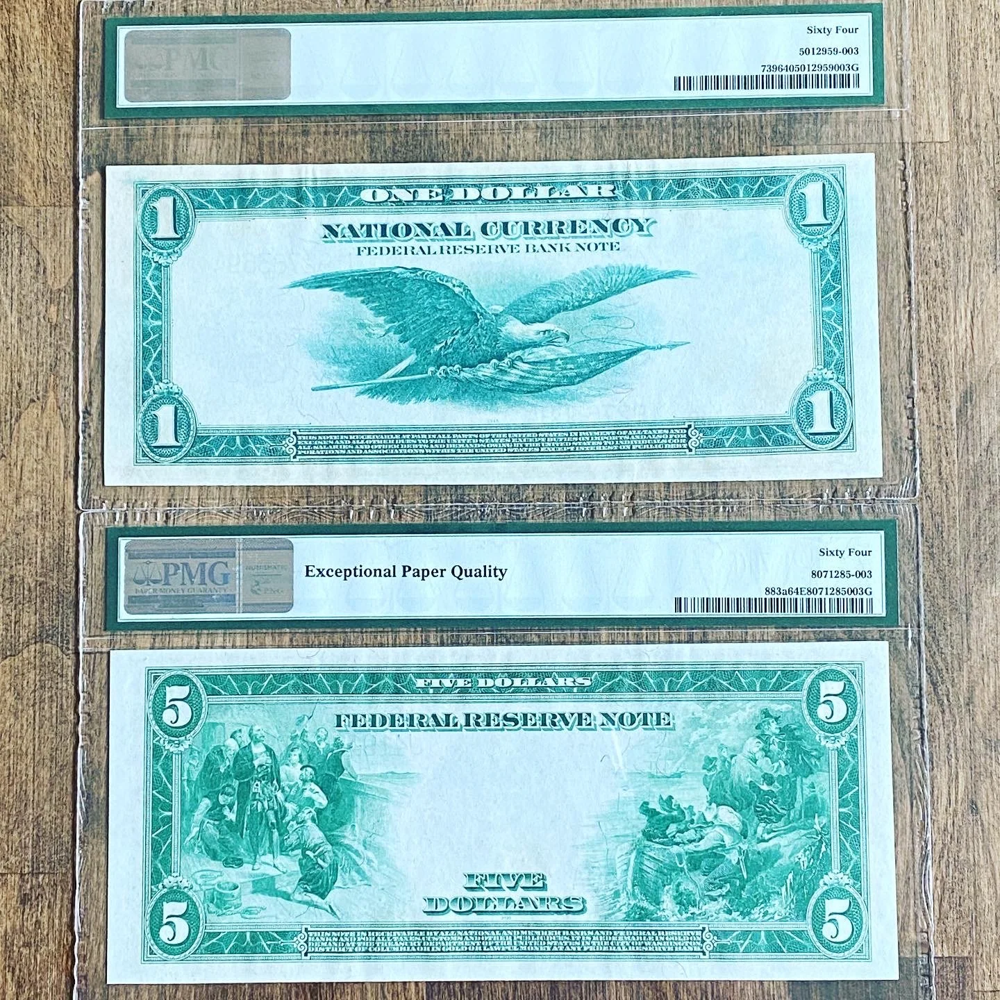 A set of three replicas of U.S. currency notes: a one-dollar bill, a five-dollar bill, and a service note, placed on a wooden surface, with a protective plastic case around the dollar bill.