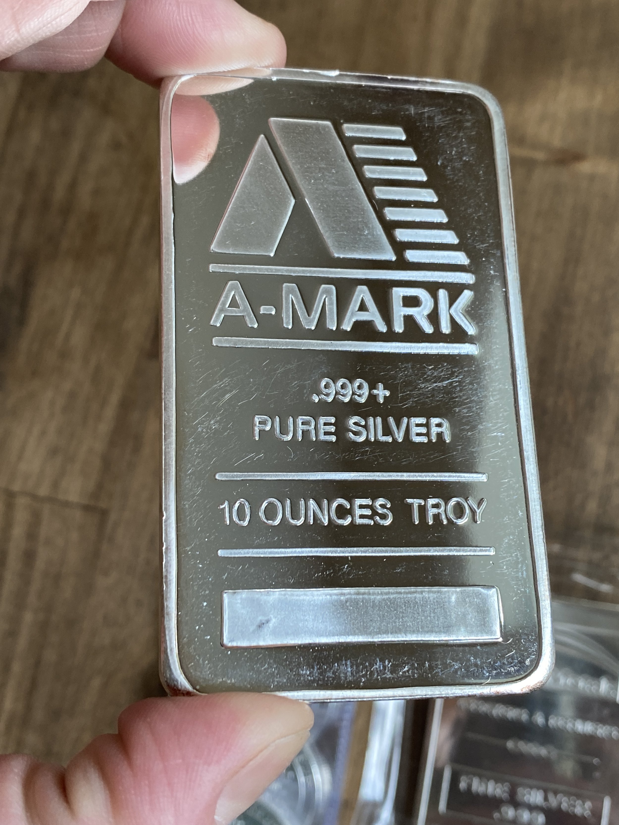 A hand holding a silver-colored rectangular metal bar with engraved text and symbols, including ANA-MARK, .999+ PURE SILVER, 10 OUNCES TROY, and a logo with a stylized triangle and lines.