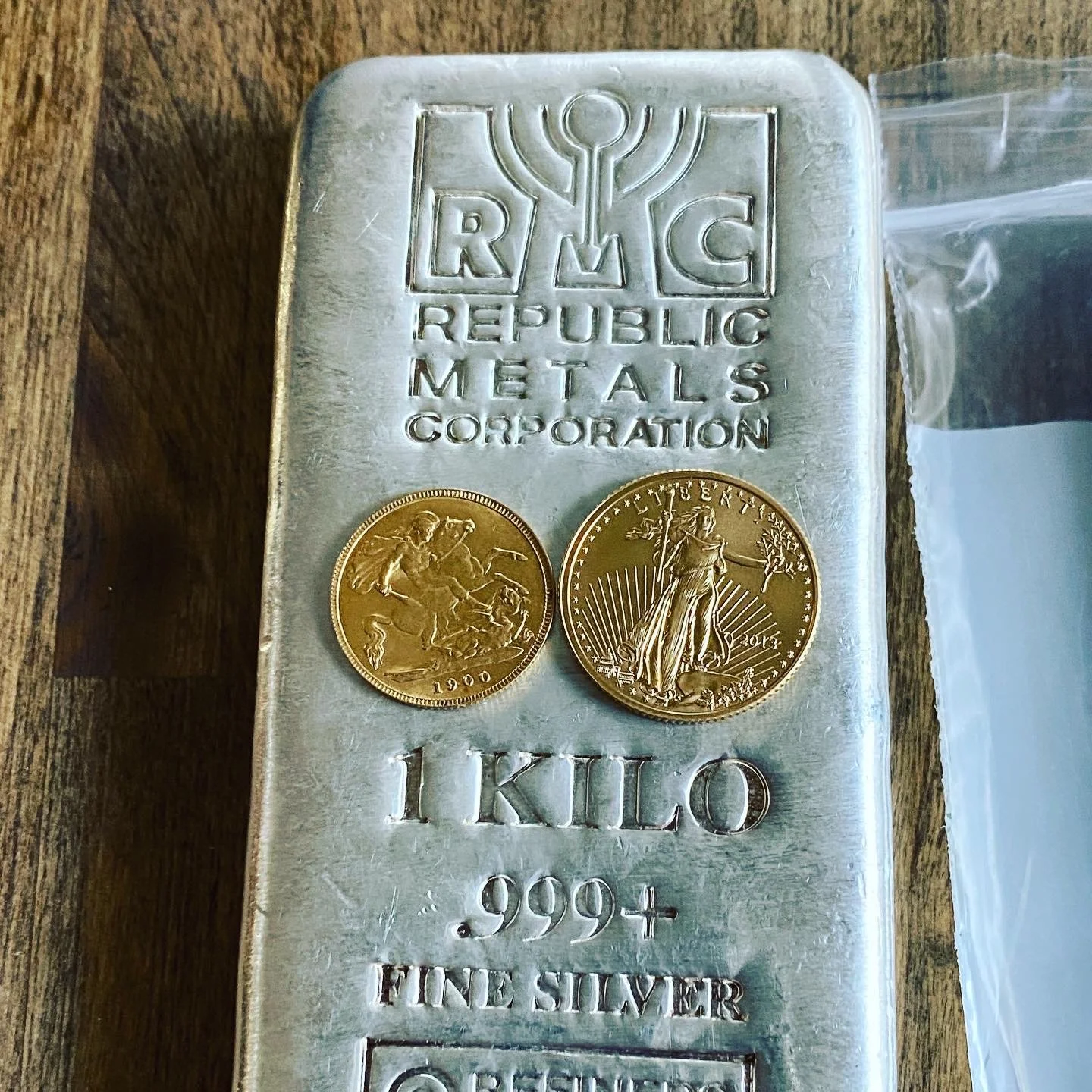 A silver bullion bar with the inscription 'RMC REPLUBLIC METALS CORPORATION 1 KILO .9994 FINE SILVER' and the RMC logo at the top. Two gold coins are placed on the bar, one showing a warrior on horseback and the year 1900, and the other showing Lady 