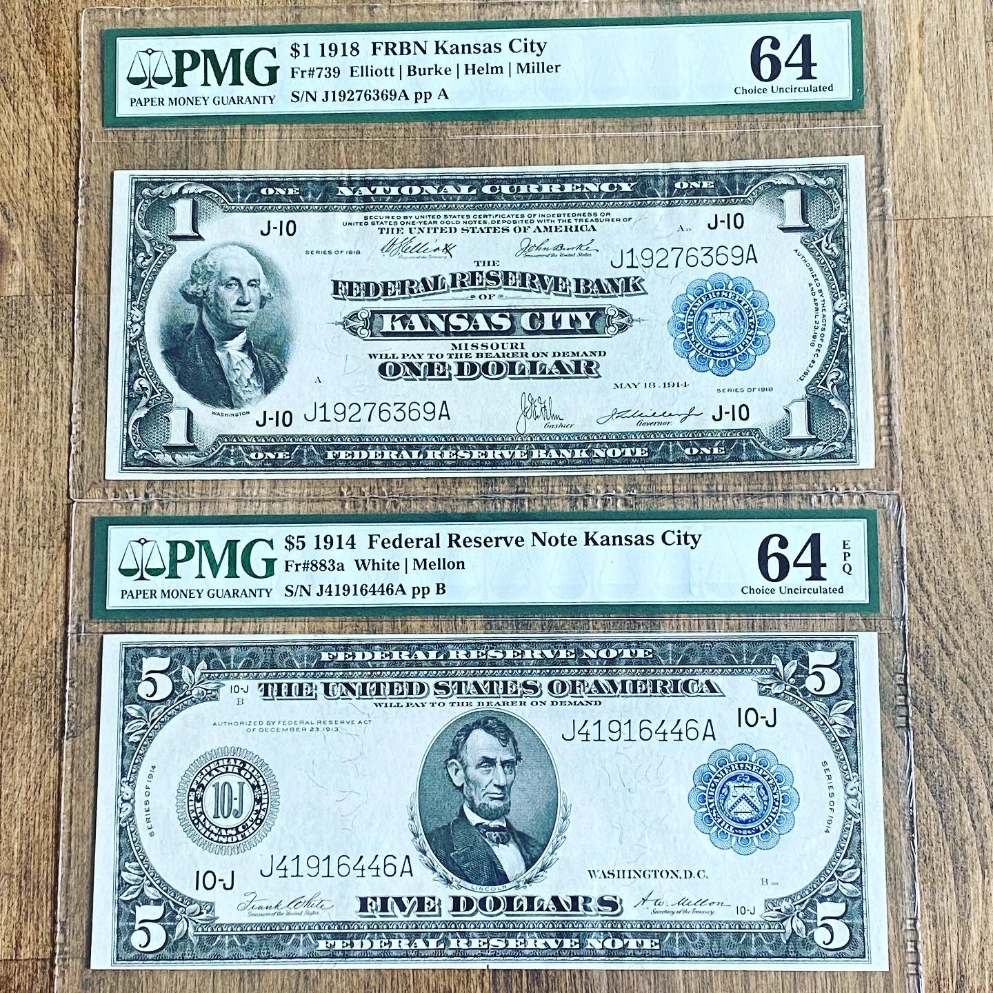 Two Kansas City Federal Reserve notes from 1914 and 1918, along with two uncirculated $1 and $5 bills sealed in protective sleeves, displayed on a wooden surface.