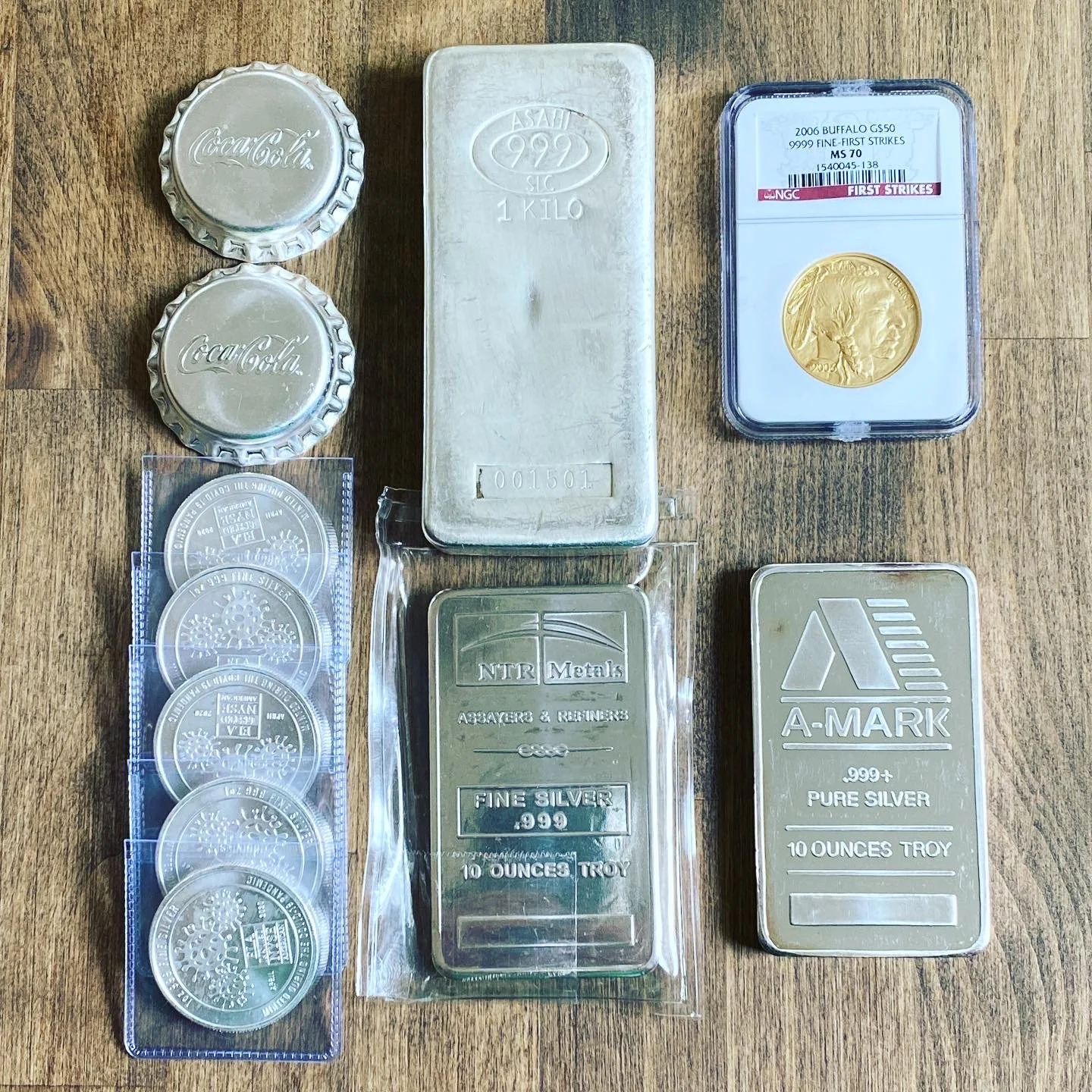 Collection of silver and gold items on a wooden surface, including two Coca-Cola bottle caps, a silver bar, a gold coin in a protective case, a group of silver coins, and two silver bars, one labeled 'NTR Metals' and the other 'A-Mark'.