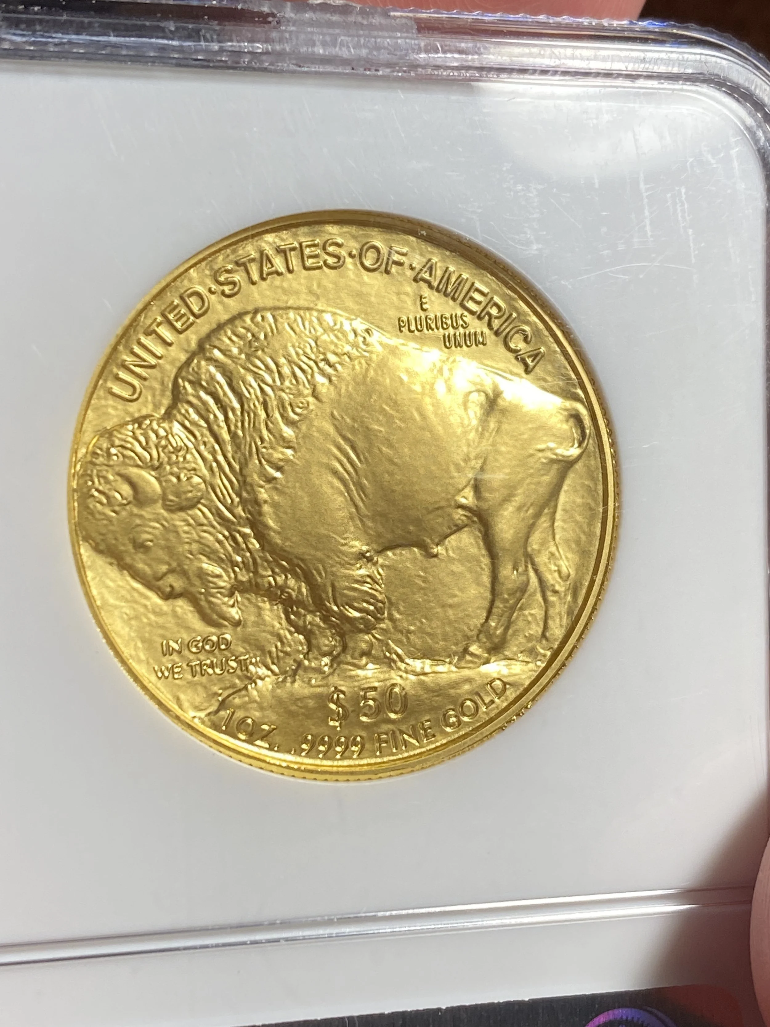 Close-up of a gold coin with a bison image, inscriptions including "United States of America," "$50," and "1 oz. .9999 fine gold," inside a plastic holder.