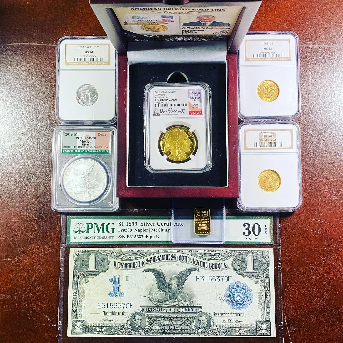 Collection of coins, a silver certificate, and a gold bar displayed on a wooden surface. The coins include a 2004 American Buffalo $25, a 2016 Mexican coin in a protective case, a 1995 $5 gold coin, and other gold coins. The silver certificate is a o