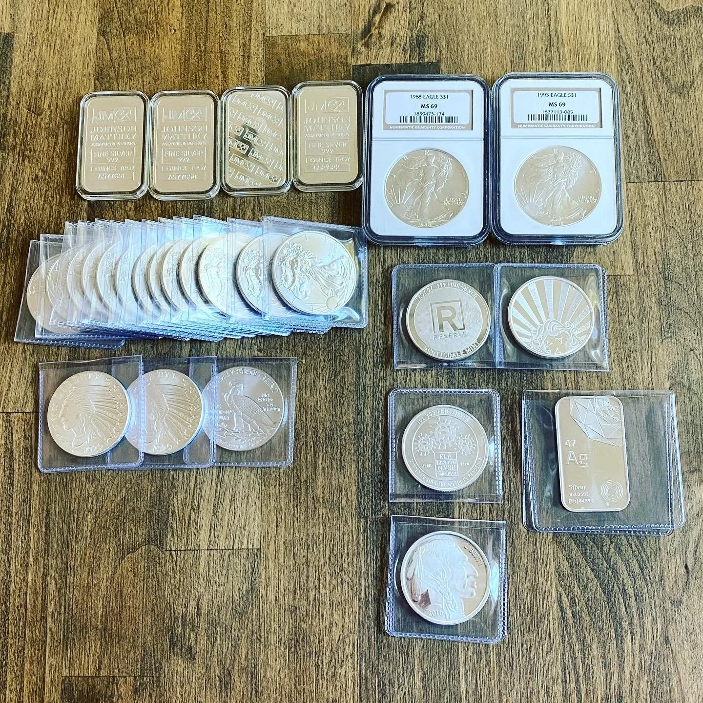 Collection of silver coins and bars displayed on a wooden surface, including various sizes and designs, some in protective cases and plastic sleeves.