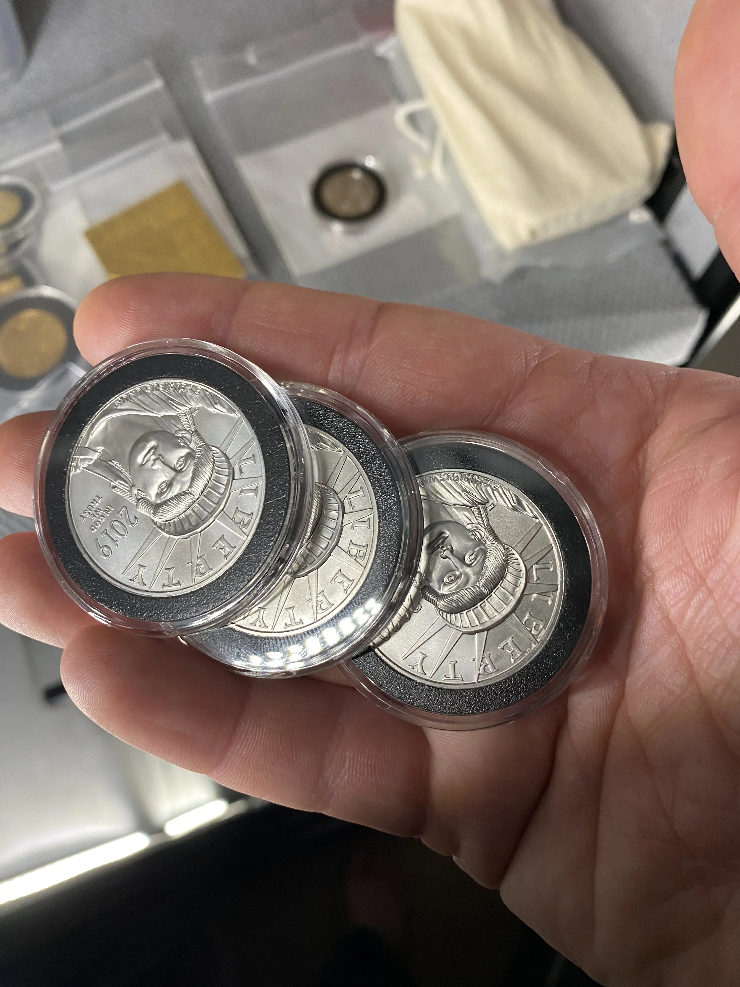 Close-up of three silver-colored commemorative coins featuring Queen Elizabeth II's profile, each encased in protective plastic cases, held in a person's hand.