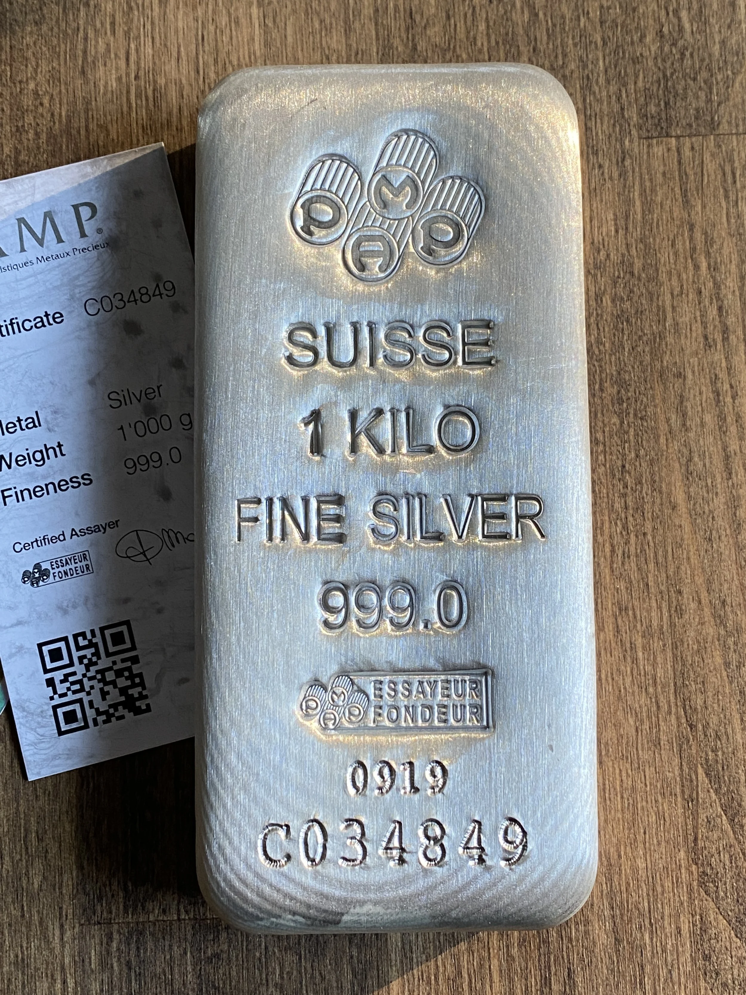Silver 1 kilogram bar with embossed text reading 'Suisse 1 Kilo Fine Silver 999.0' and a logo of five silver coins above the text. An official certificate paper is partially visible beside the bar.