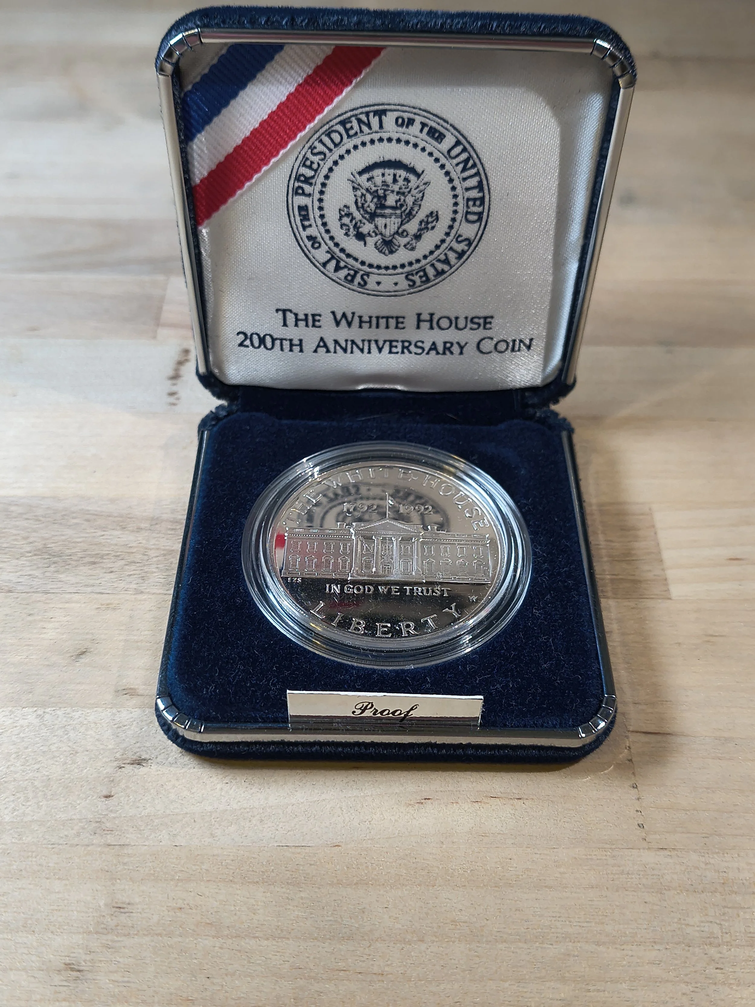 A silver coin in a presentation case with a navy blue interior, commemorating the 200th anniversary of the White House, featuring an engraved image of the White House and the words "In God We Trust" and "Liberty." The case has a label that says "Proo