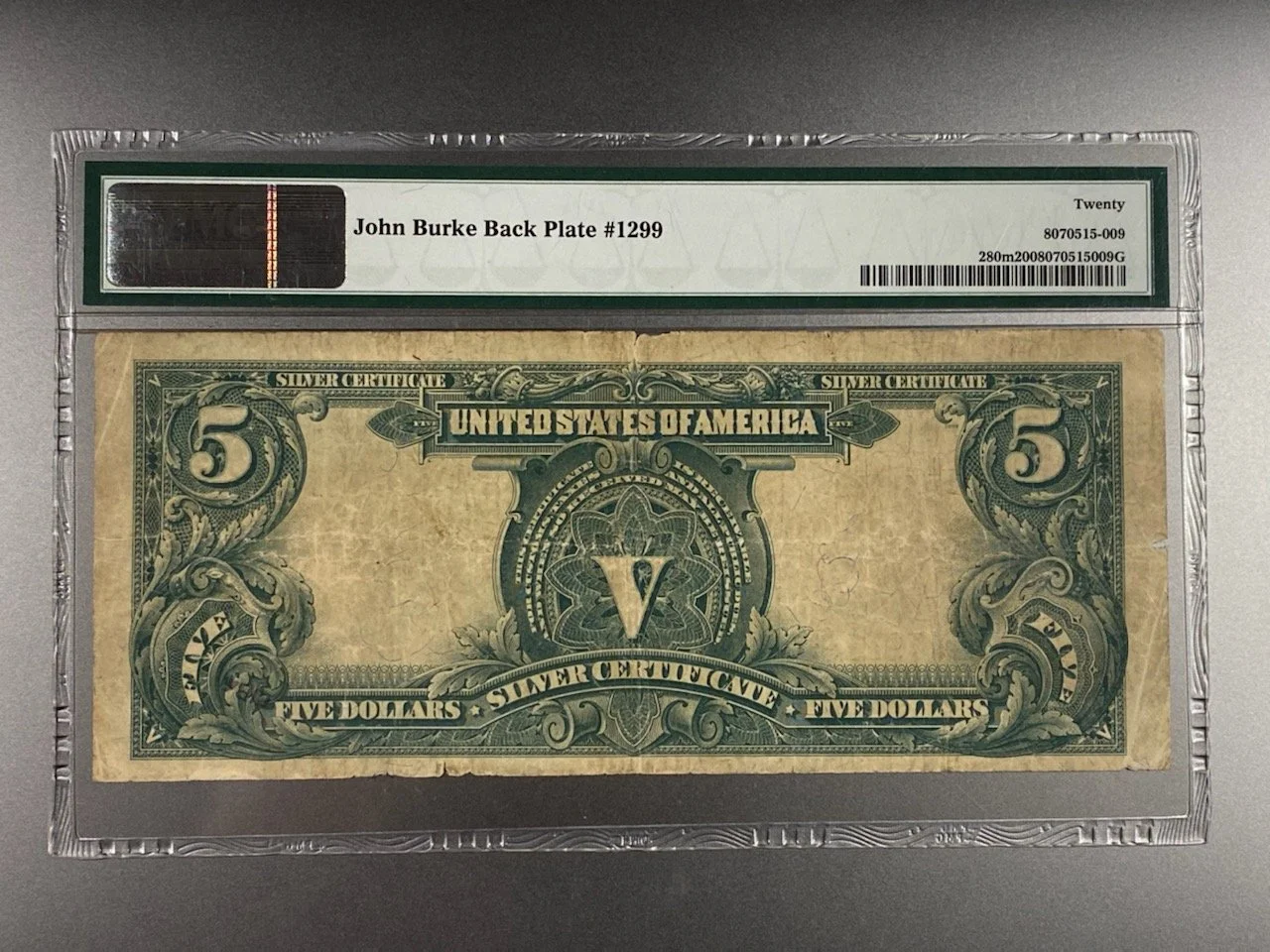 A framed, vintage five dollar bill with intricate green and black designs, labeled 'United States of America'. The bill is encased in a plastic holder with a label at the top reading 'John Burke Back Plate #1299'.
