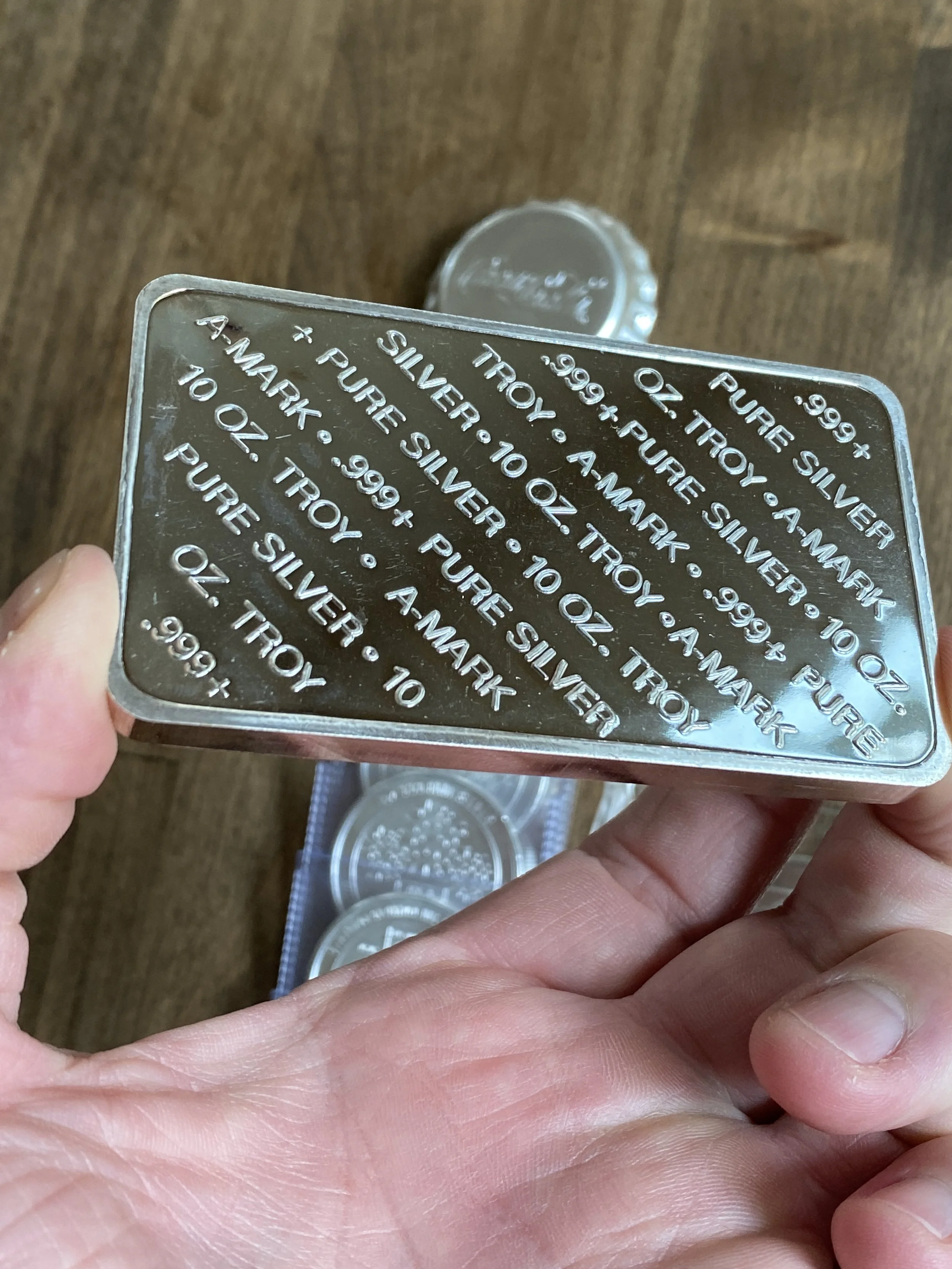 A person holding a silver bar with engraved text, and several coins behind it on a wooden surface.