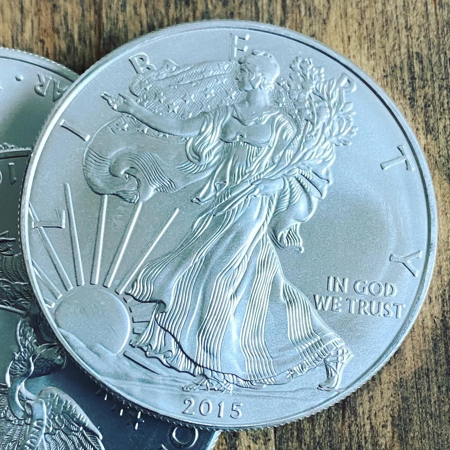 A silver coin featuring Lady Liberty walking with a staff and olive branch, with rays of sunlight and the words "LIBERTY" and "IN GOD WE TRUST." The coin is dated 2015.
