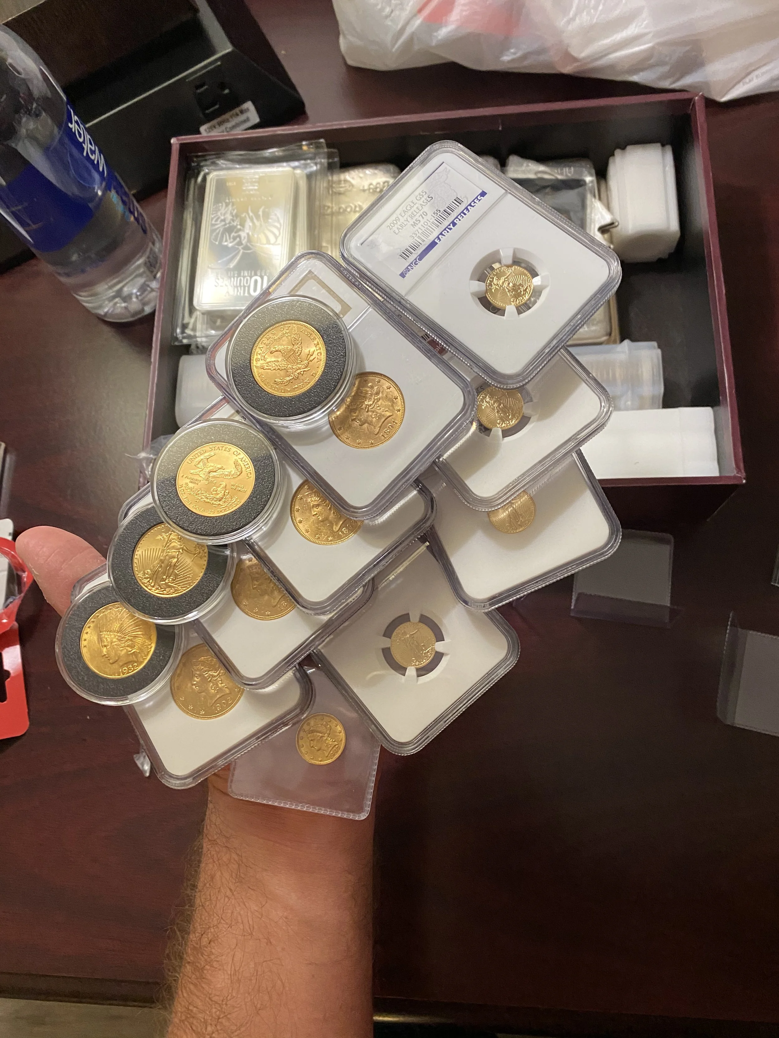 A person's hand holding multiple gold coins in protective plastic cases, with a water bottle and a wooden surface with some boxes and packaging in the background.
