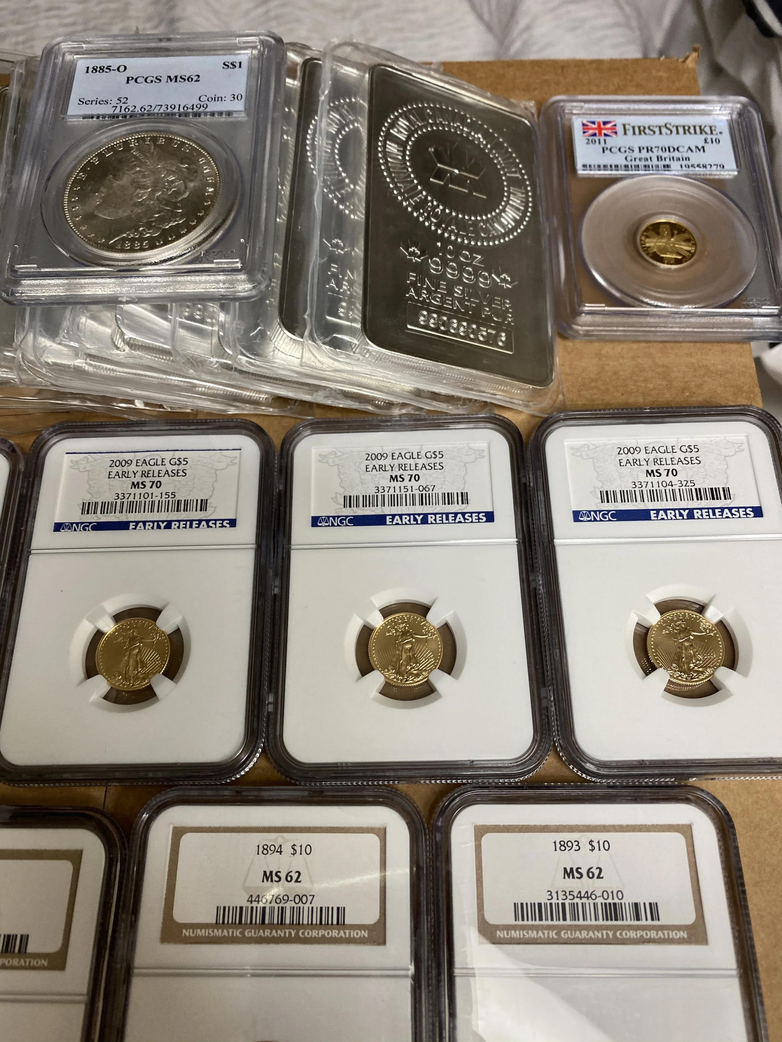 A collection of graded and ungraded coins, including a 1894 and 1893 silver dollar, a 2009 gold eagle coin, an 1885 Morgan silver dollar, and a silver bar, all organized in protective cases.