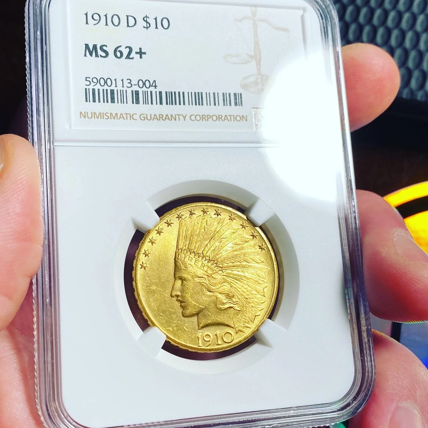 A 1910 D $10 gold coin in a protective plastic case labeled MS 62+. The coin features a Lady Liberty wearing a Native American headdress, with the date 1910 at the bottom.