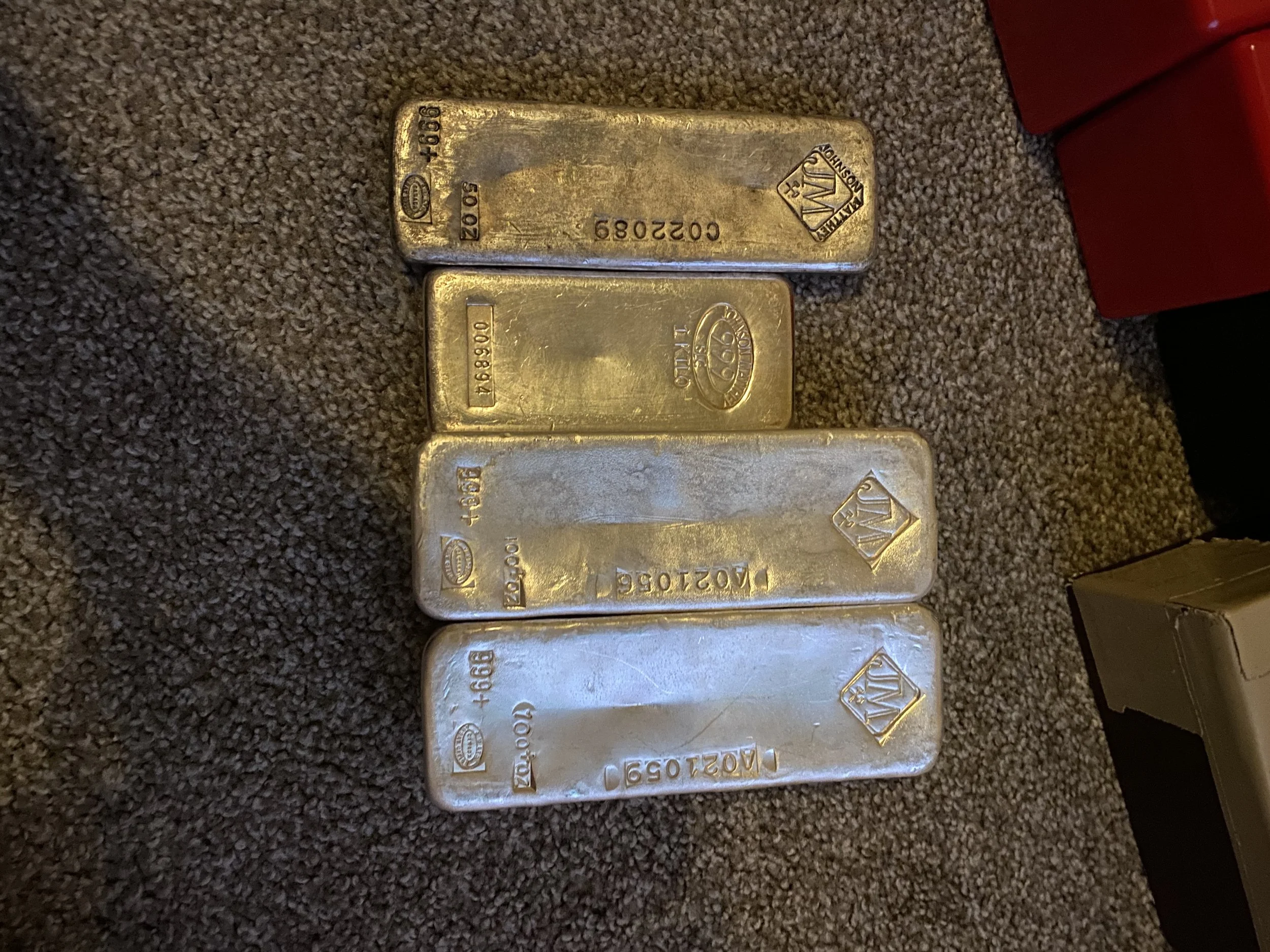 Four silver bars with engraved markings and logos, placed on a grey carpeted surface.