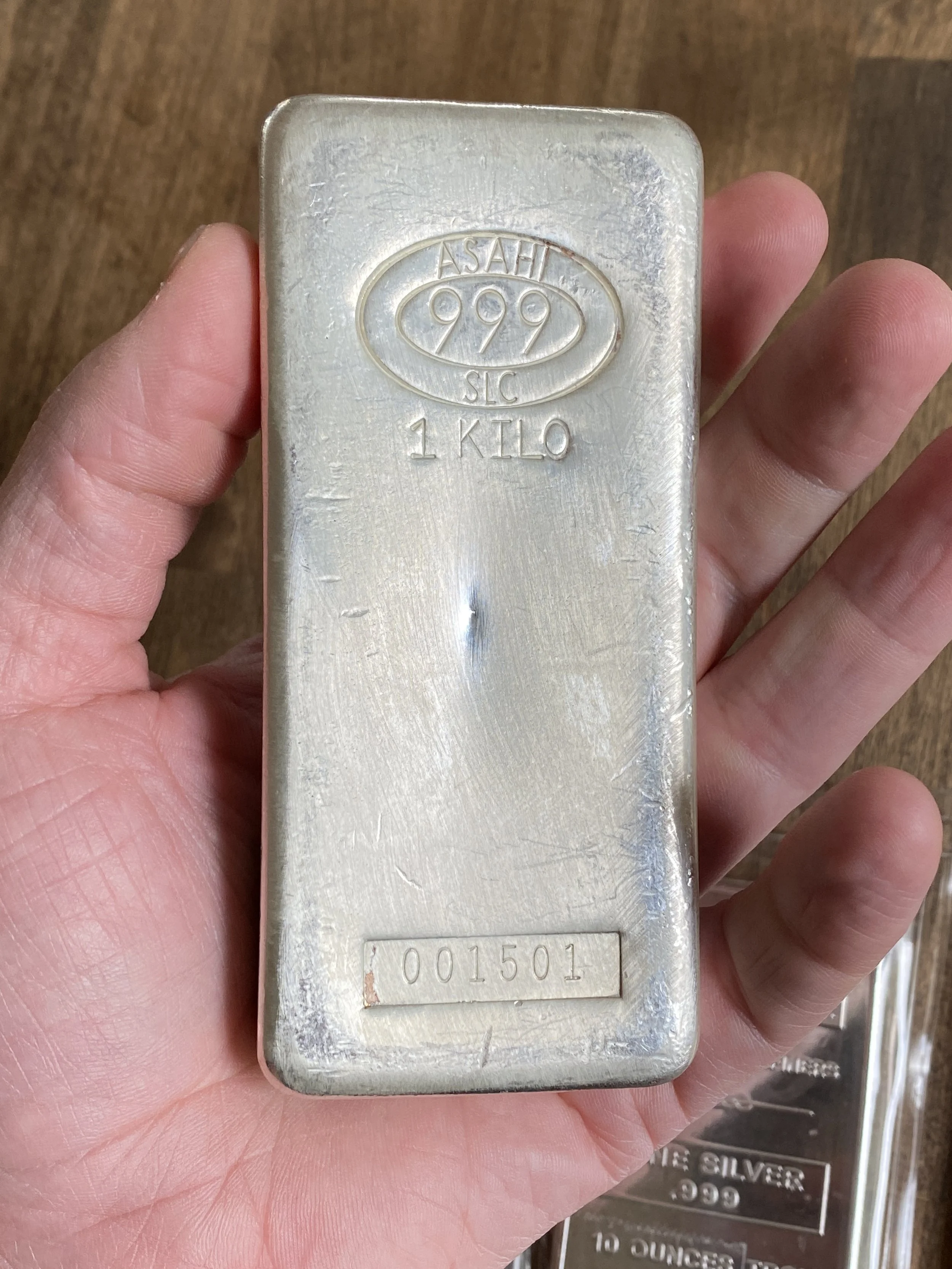 Hand holding a silver 1 kilogram silver bar with the brand 'ASAHI 999.9' and a serial number.