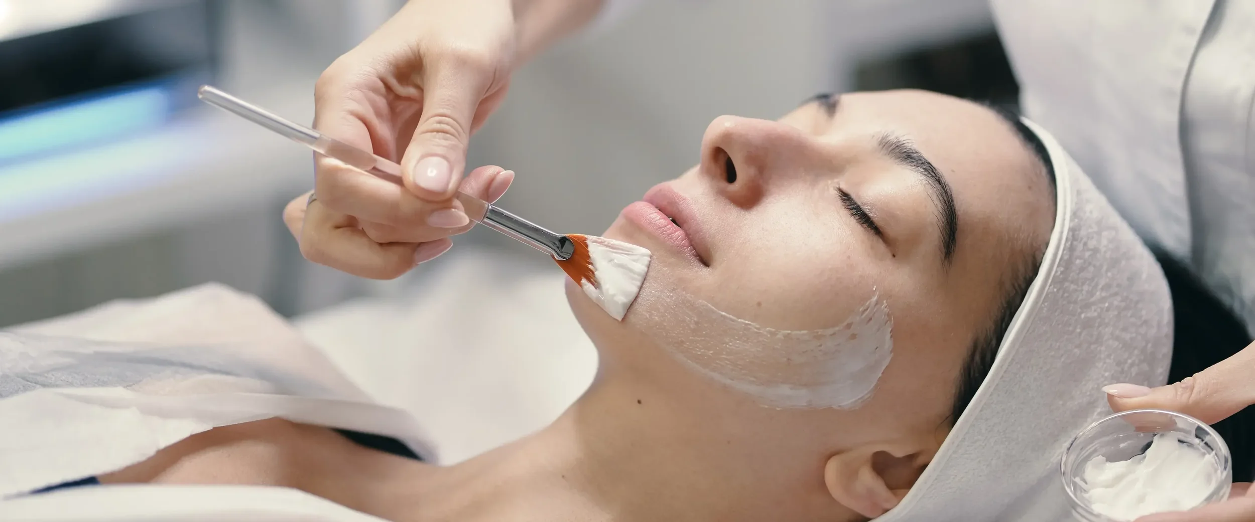 A woman receiving a facial treatment at a spa, lying with her eyes closed, while a skincare professional applies a gentle chemical peel  to her face with a small brush.