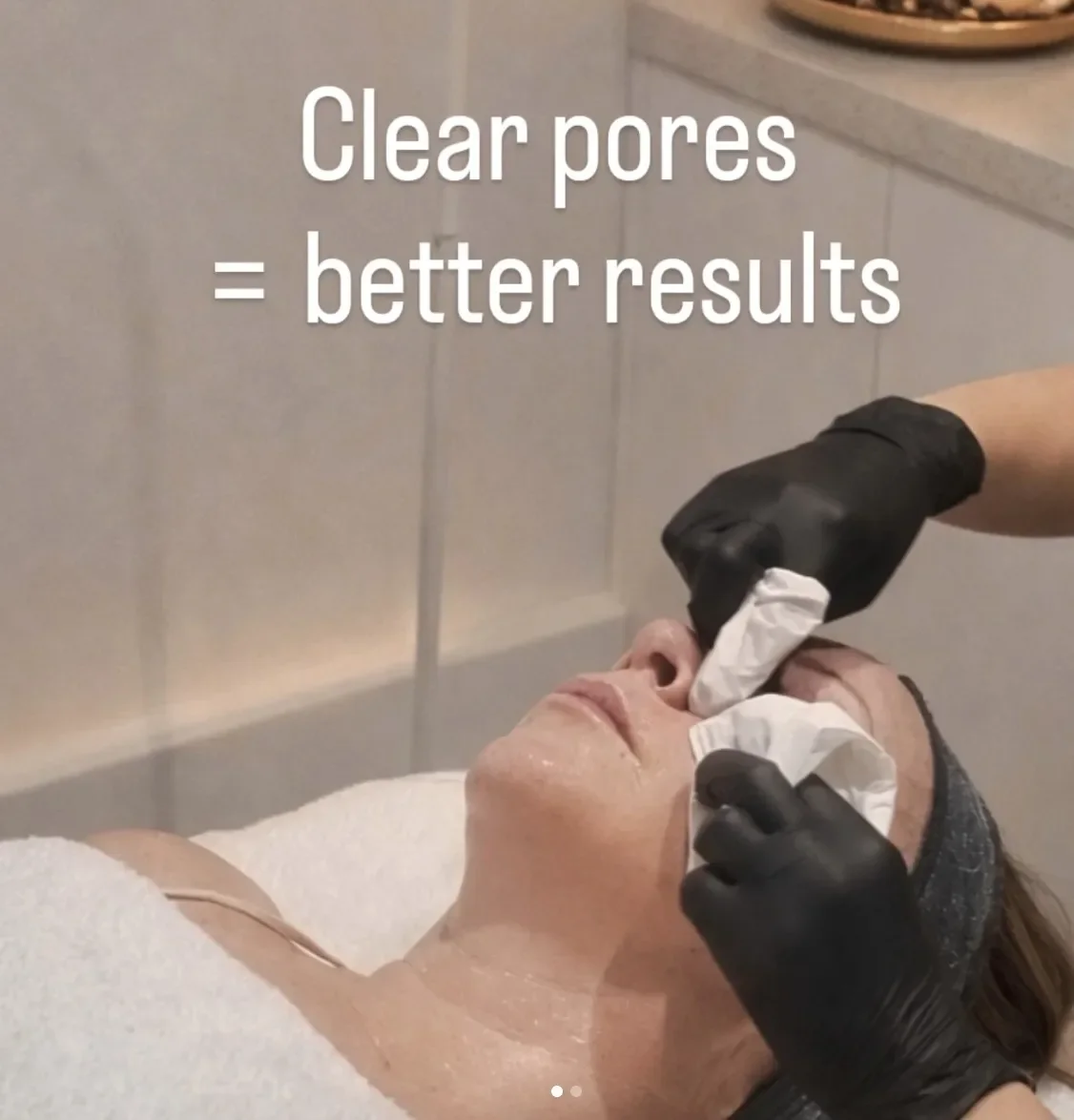 A woman receiving a facial treatment while lying down. Someone wearing black gloves is applying a cream or mask to her face with a cloth. The text overlay reads 'Clear pores = better results'.