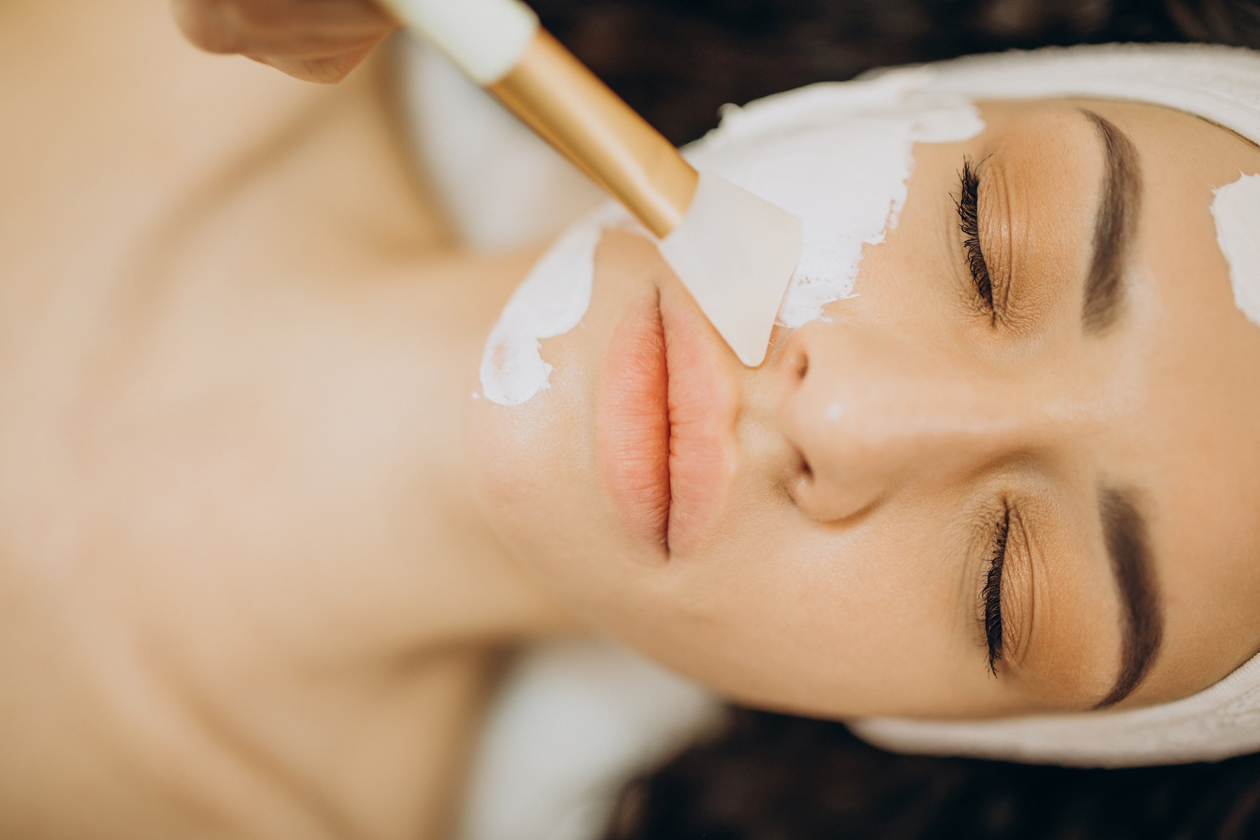 A woman receiving a facial treatment with a metal spatula applied to her face while lying down with her eyes closed, wearing a towel wrapped around her head.