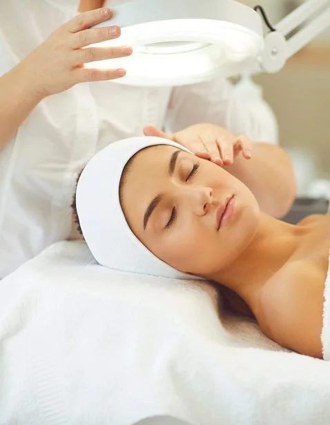 A woman receiving a facial treatment under a magnifying light in a spa or clinic setting.