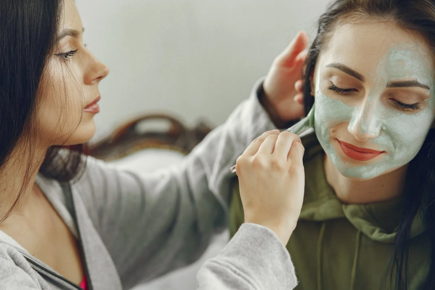A woman applies a facial mask to another woman who is sitting with her eyes closed.